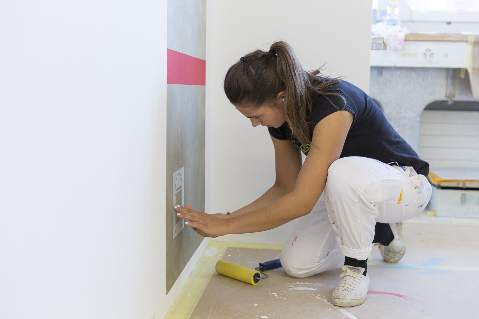Eine Frau in weißer Hose und schwarzem Hemd kniet auf dem Boden und streicht oder klebt sorgfältig in der Nähe einer Steckdose an der Wand, während sie eine Farbrolle und andere Werkzeuge in der Nähe hat.
