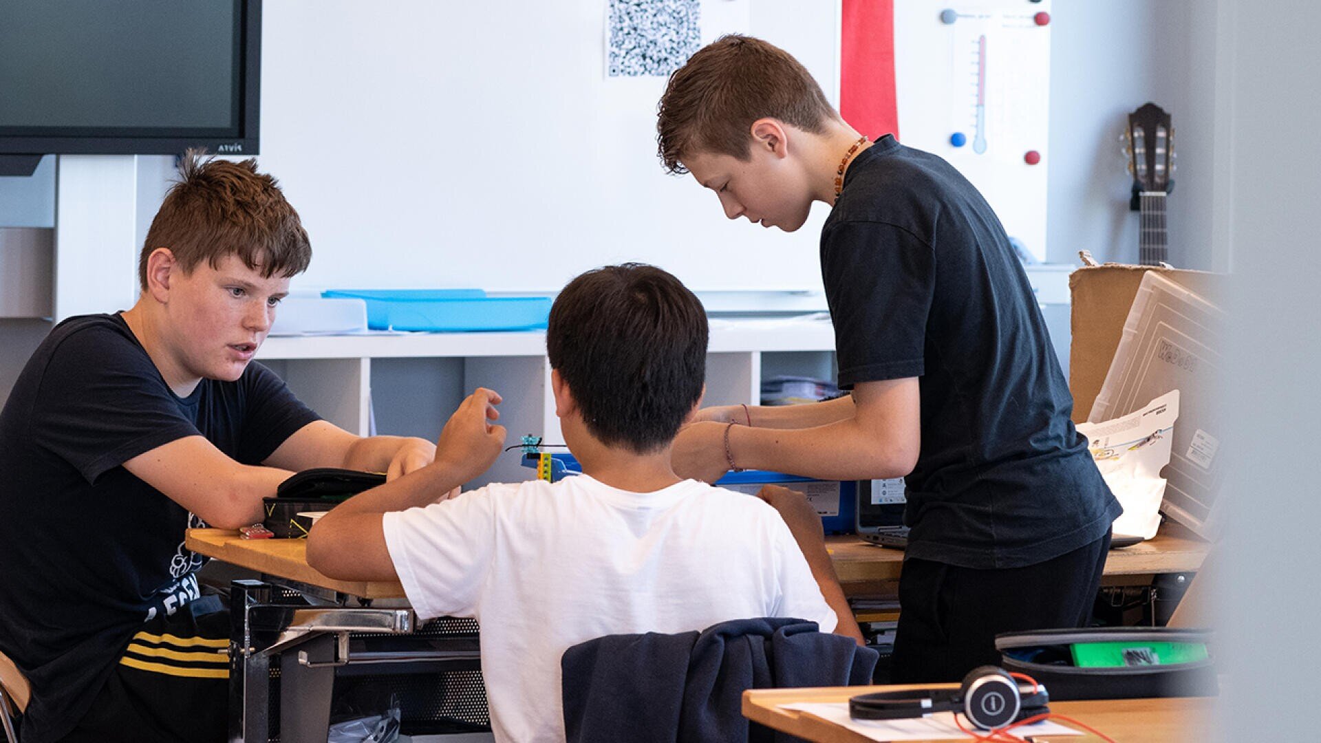 Drei Jungen sitzen und stehen um einen Tisch in einem Klassenzimmer und arbeiten gemeinsam an einer Aufgabe. Ein Junge spricht, ein anderer lehnt sich über den Tisch, und der dritte sitzt mit dem Rücken zur Kamera. Klassenzimmerzubehör ist zu sehen.