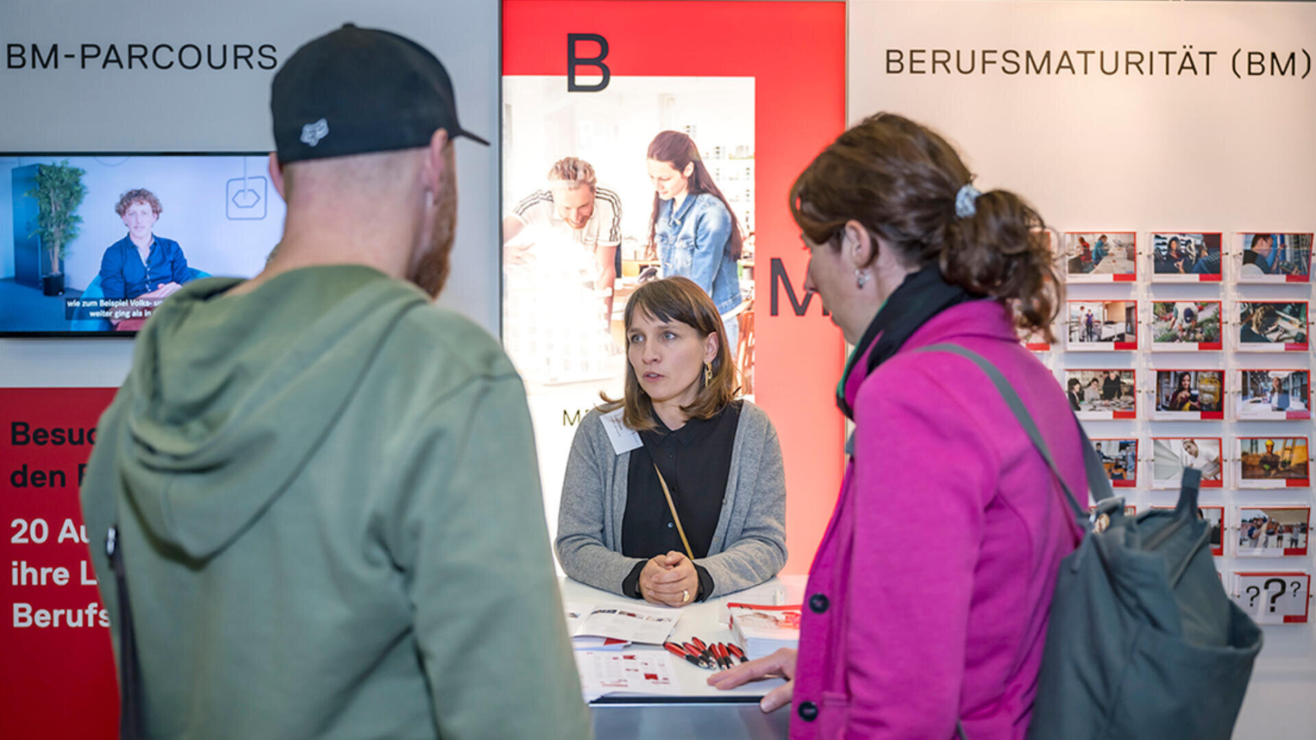 Eine Frau sitzt an einem Informationsstand mit der Aufschrift Berufsmaturität (BM) und unterhält sich mit einem Mann mit einer Mütze und einer Frau in einer rosa Jacke auf einer Bildungsmesse. Auf dem Tisch und an den Wänden sind Plakate und Broschüren zu sehen.