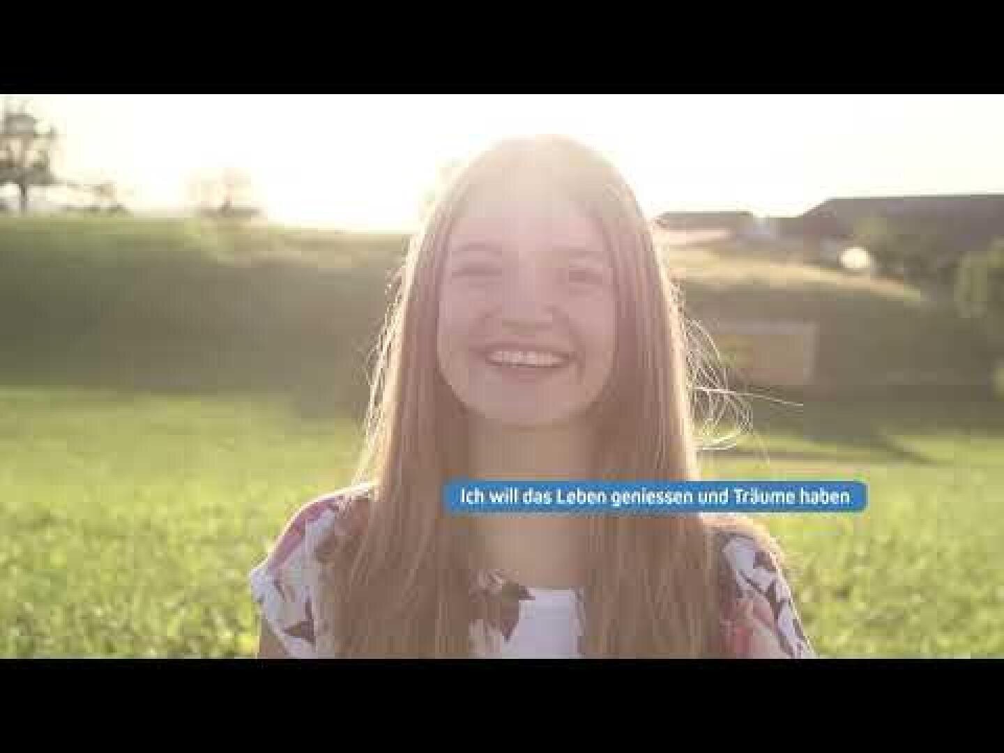 Ein lächelndes Mädchen steht draußen auf einer sonnenbeschienenen Wiese. Das Sonnenlicht scheint hinter ihr und schafft eine warme, helle Atmosphäre. In einem blauen Textfeld steht: "Ich will das Leben genießen und Träume haben.