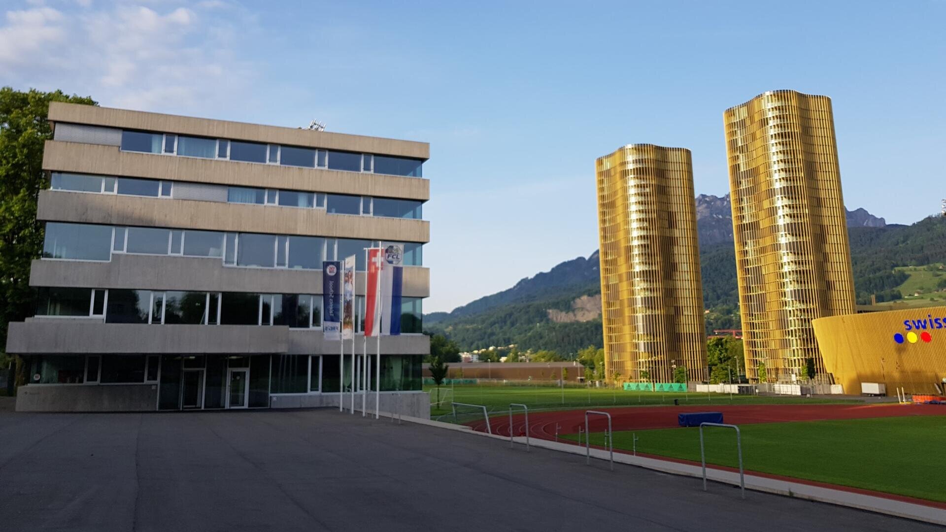 Ein modernes graues Gebäude steht neben einem grünen Sportplatz und einer Laufbahn, mit zwei hohen, goldglänzenden Türmen und Bergen im Hintergrund unter einem klaren blauen Himmel.
