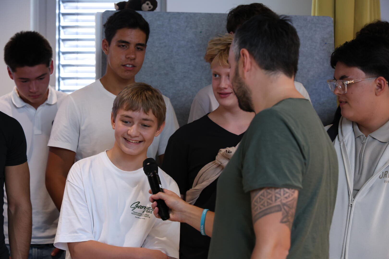 Ein Mann hält ein Mikrofon in Richtung eines lächelnden Jungen in einer Gruppe von Teenagern. Die Gruppe steht in einem Haus, leger gekleidet, einige schauen den Jungen an, andere schauen nach unten.