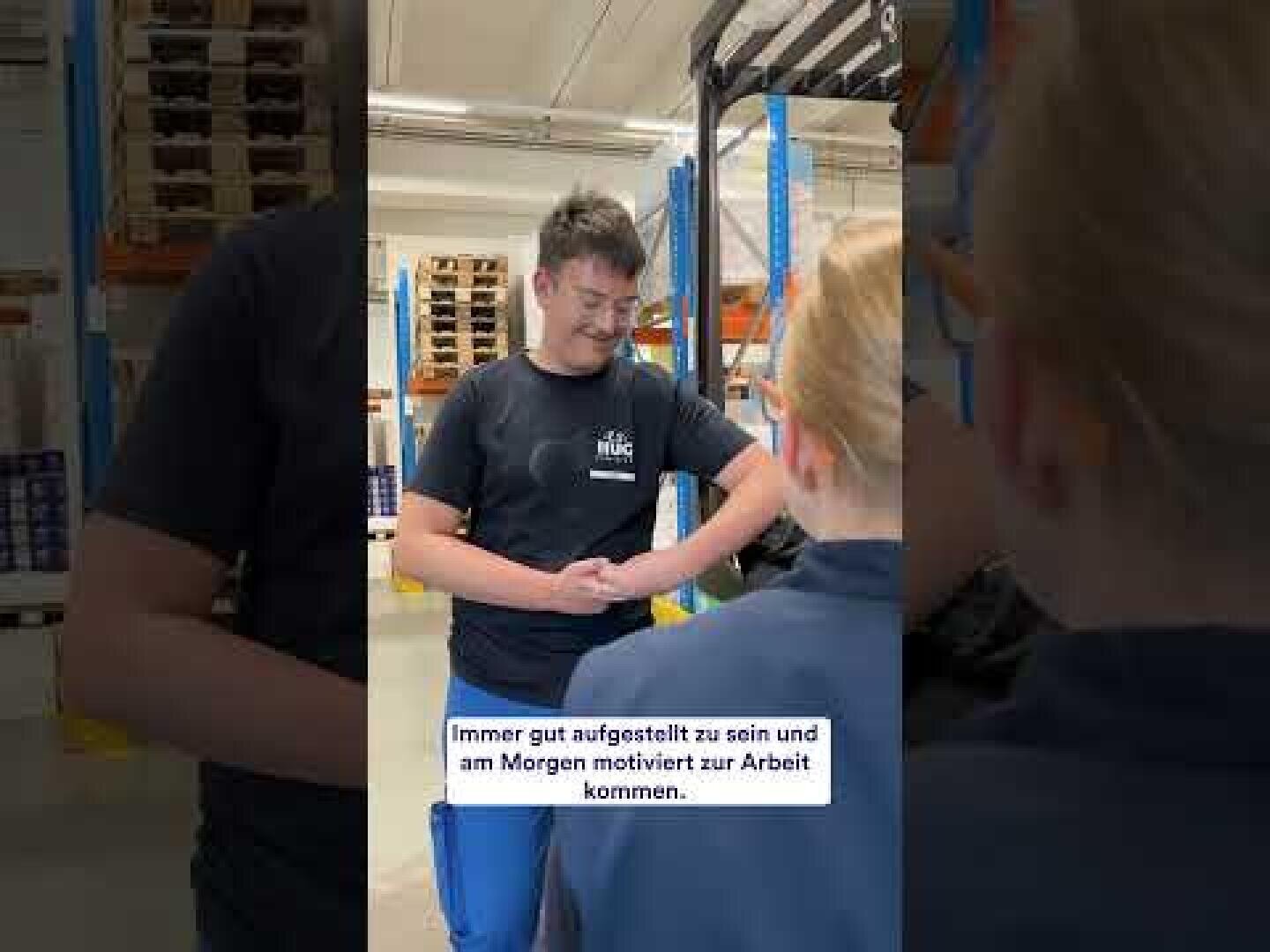 Ein junger Mann in einem Lagerhaus lächelt, während er seinen Ärmel hochkrempelt und einen Mitarbeiter ansieht. Der deutsche Text auf dem Bild lautet: Immer gut aufgestellt zu sein und am Morgen motiviert zur Arbeit zu kommen.
