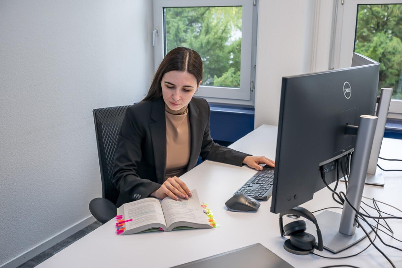 Eine Frau in einem Blazer sitzt an einem Schreibtisch und blickt auf ein aufgeschlagenes Lehrbuch mit bunten Lesezeichen.