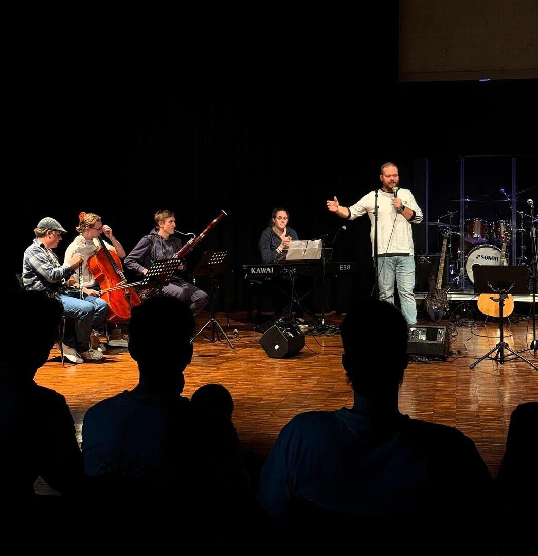 Auf der Bühne steht ein Mann und spricht in ein Mikrofon, neben ihm spielen Musiker Cello, Klarinette, Keyboard und Gitarre, während das Publikum in einem schwach beleuchteten Raum zusieht.