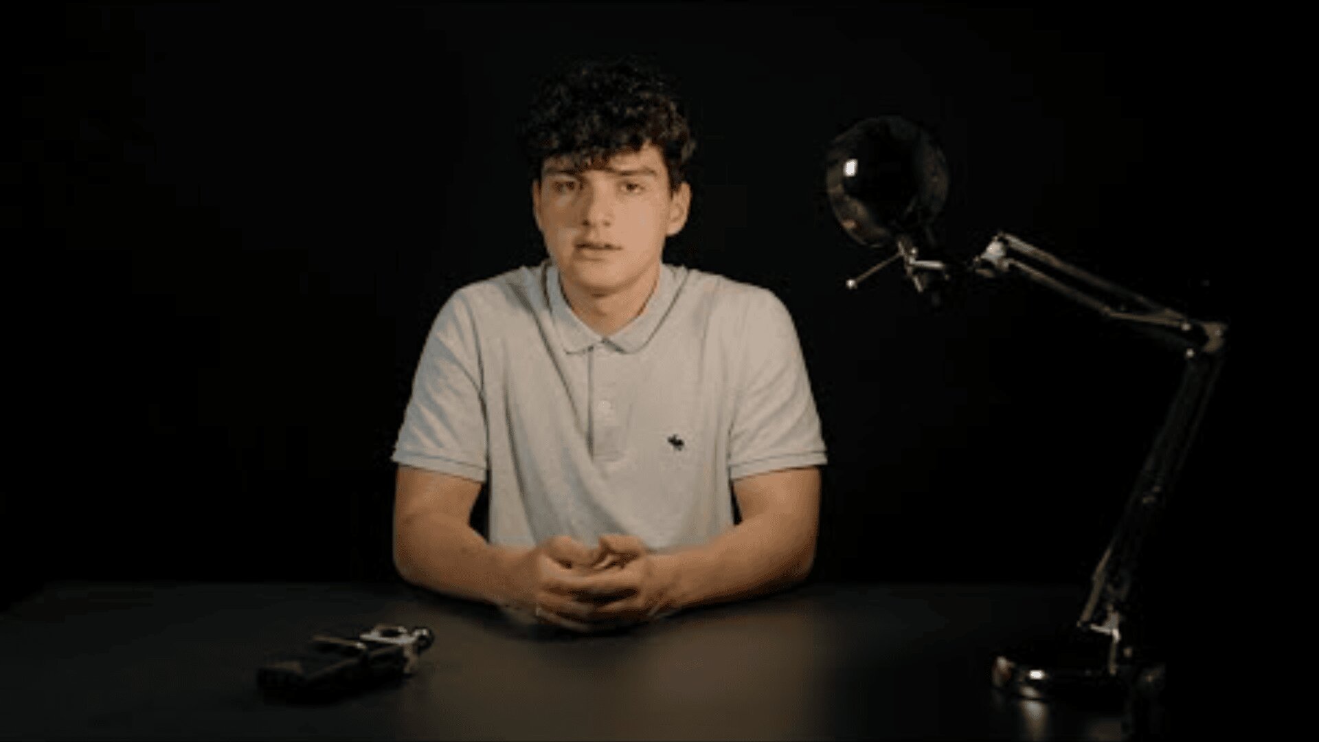 Ein junger Mann mit lockigem dunklem Haar sitzt an einem dunklen Tisch und trägt ein helles Poloshirt. Auf dem Tisch neben ihm befinden sich eine Schreibtischlampe und eine Handfeuerwaffe, alles vor einem schwarzen Hintergrund.