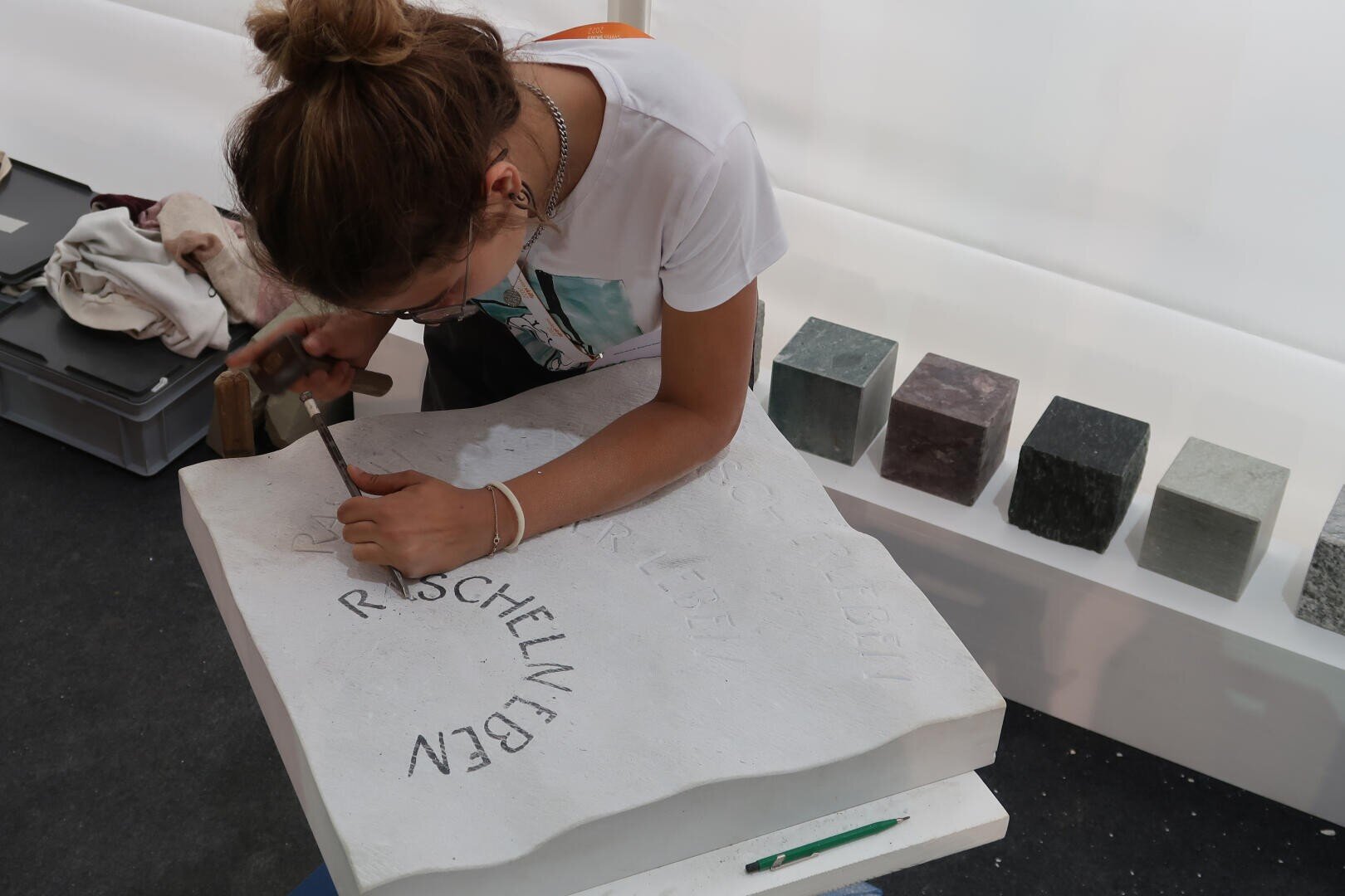 Eine Person mit zusammengebundenen Haaren meißelt mit einem Meißel Buchstaben in eine große weiße Steinplatte. Im Hintergrund sind auf einem weißen Regal mehrere Steinblöcke zu sehen.