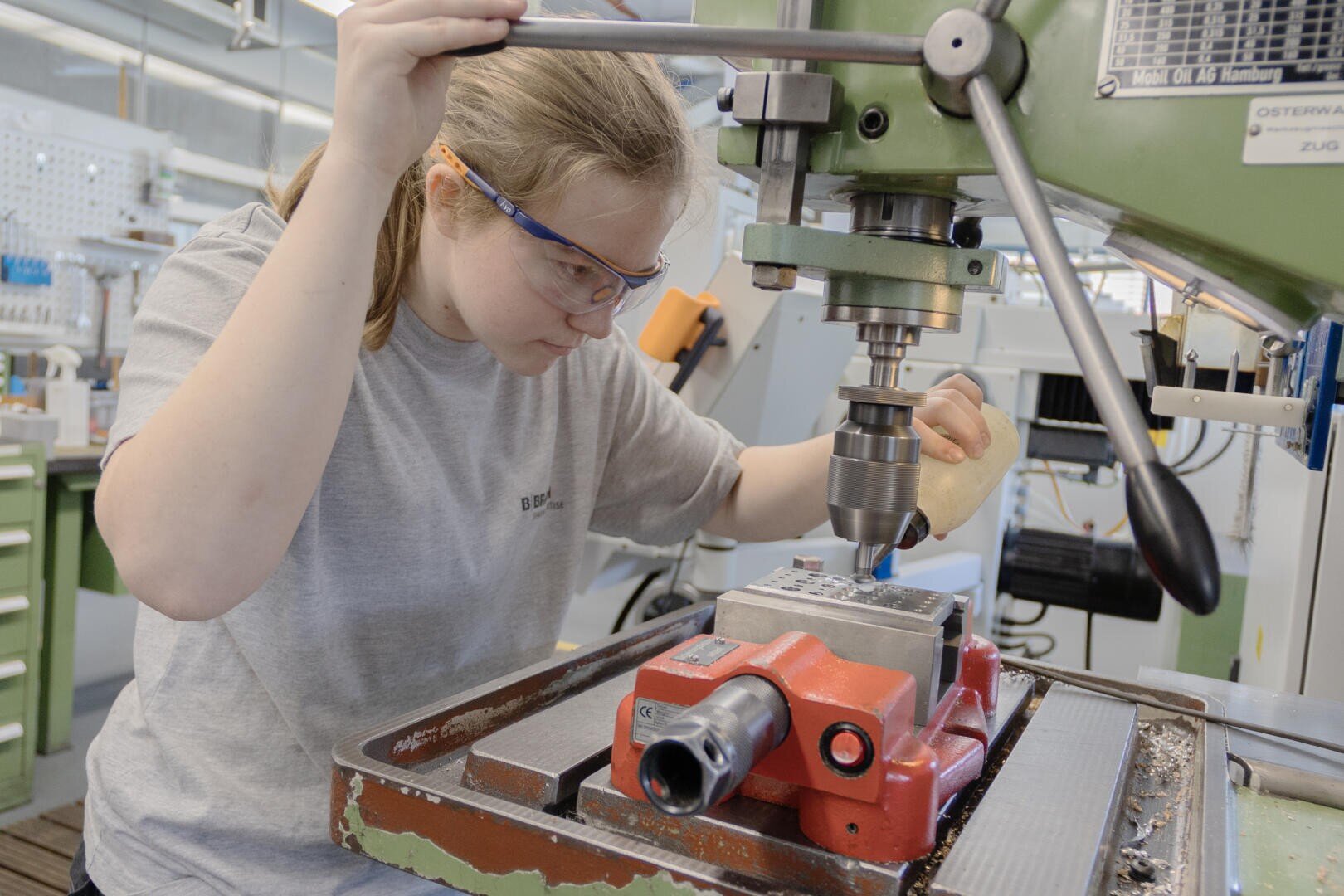 Eine Person, die eine Schutzbrille trägt, bedient in einer Werkstatt eine Bohrmaschine und konzentriert sich darauf, ein Werkstück zu bohren, das in einem roten Schraubstock eingespannt ist.