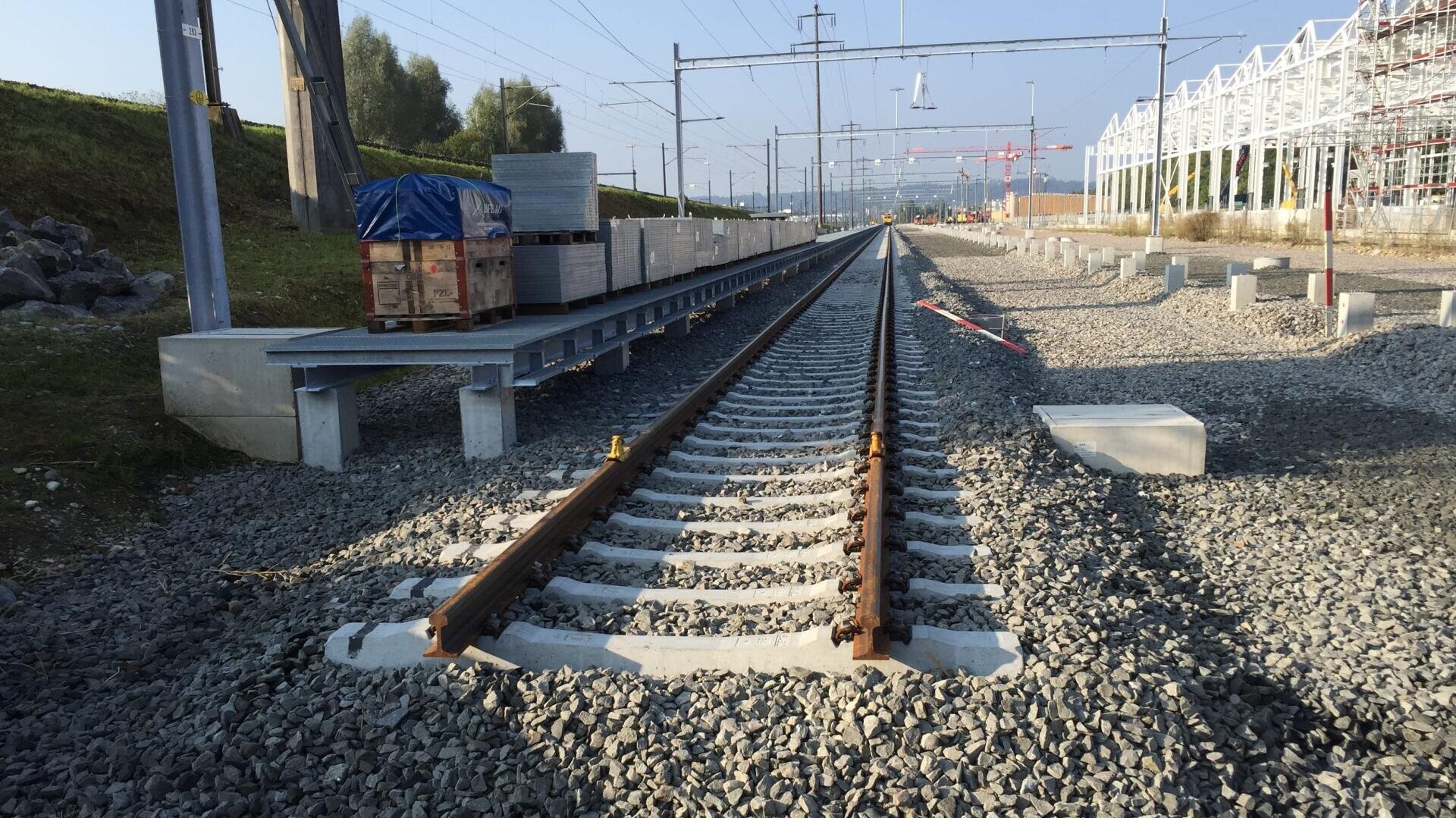 Eisenbahnschienen im Bau, umgeben von Schotter. Links stapeln sich die Baumaterialien, rechts erhebt sich eine weiße Metallkonstruktion. Oberleitungen und Masten verlaufen oberhalb der Gleise in der Ferne.