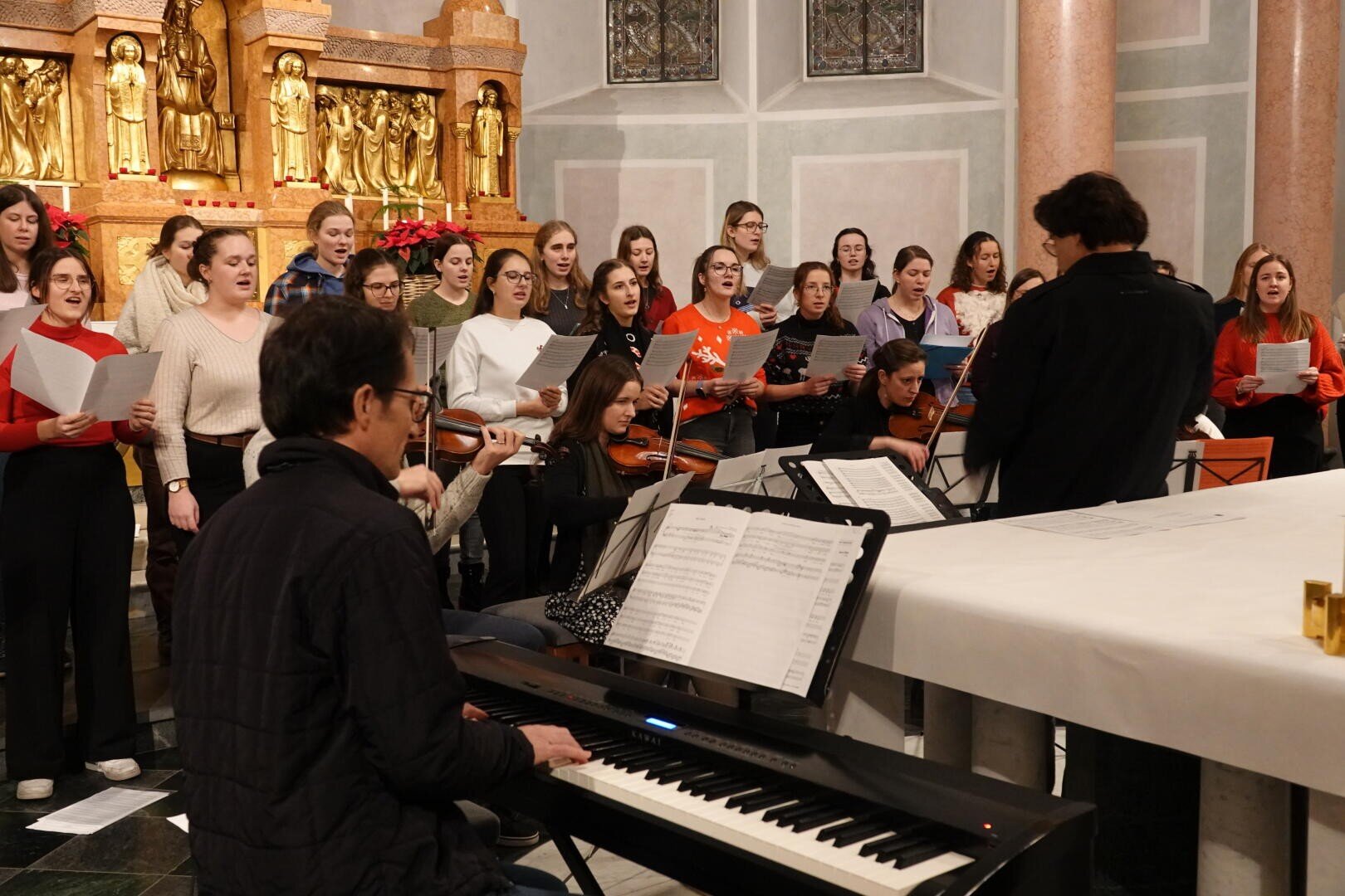 Ein Chor und Musiker treten in einer Kirche auf. Der Chor singt unter der Leitung eines Dirigenten, und im Vordergrund spielt ein Pianist auf einem Keyboard. Im Hintergrund sind Streicher und kunstvoll verzierte goldene religiöse Statuen zu sehen.
