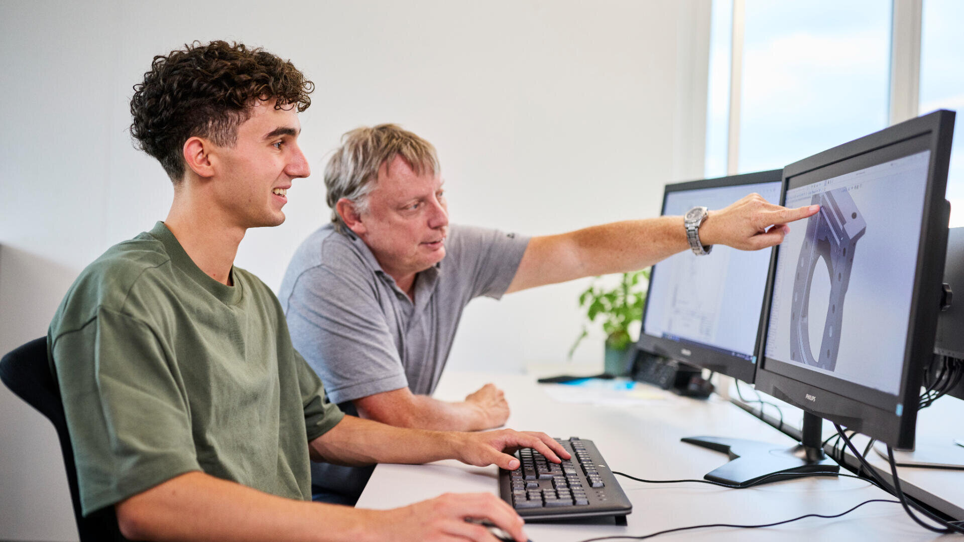 Zwei Personen sitzen an einem Schreibtisch und benutzen Computer. Einer tippt, während der andere auf eine technische Zeichnung auf dem Monitor zeigt, was auf eine Zusammenarbeit bei einem Designprojekt hindeutet. Beide sind konzentriert und beschäftigt.