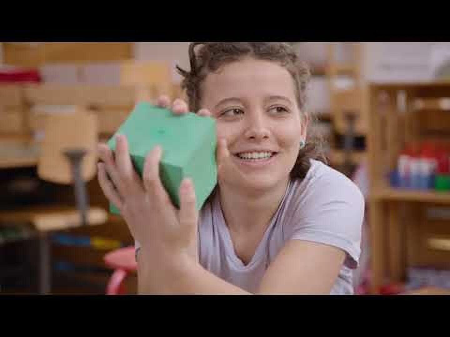 Ein lächelnder junger Mensch in einem hellen Hemd hält mit beiden Händen einen grünen Schaumstoffblock in einem Klassenzimmer, in dem im Hintergrund Regale und Material zu sehen sind.