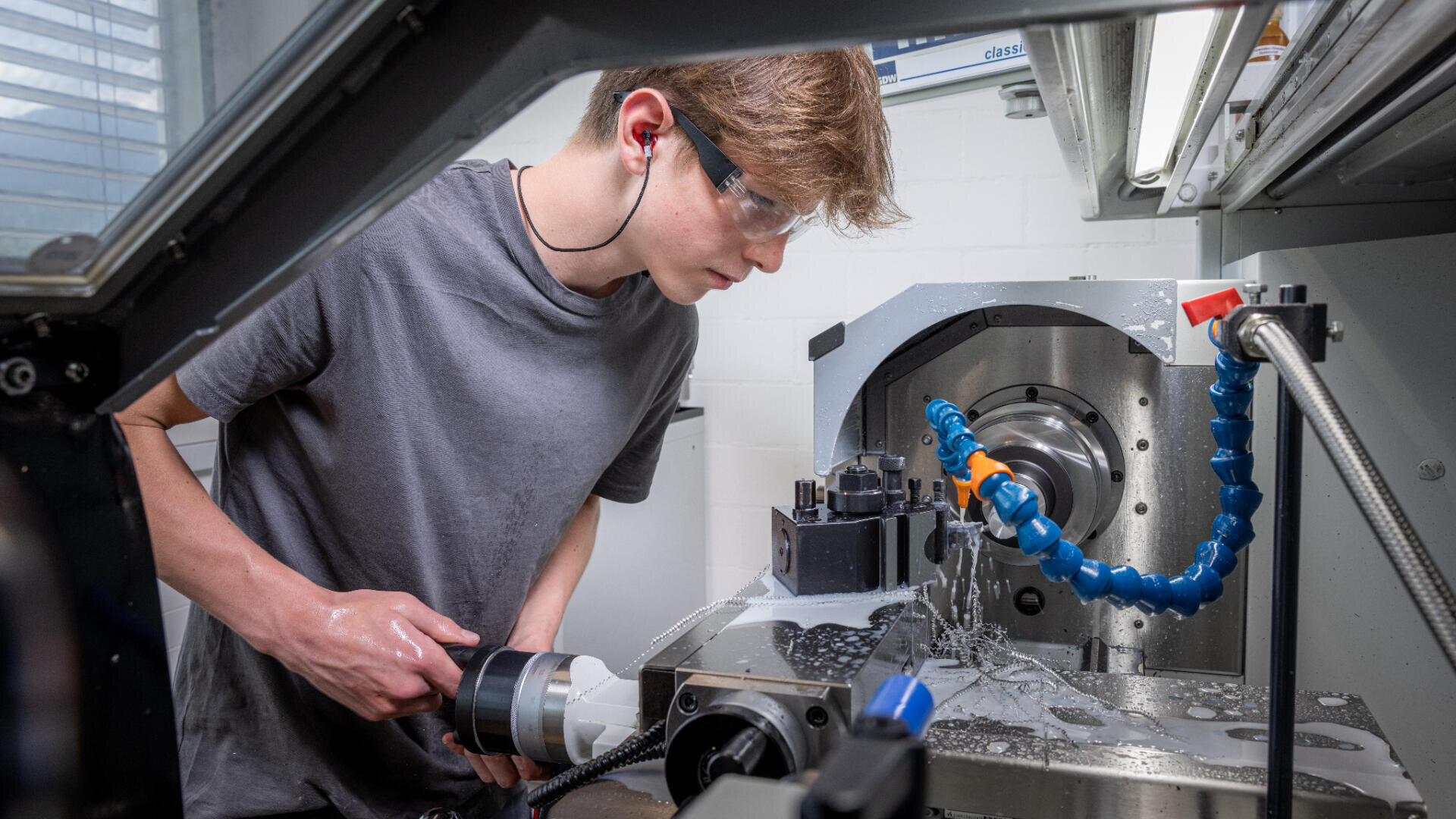 Ein Jugendlicher, der eine Schutzbrille und einen Gehörschutz trägt, bedient eine Metallbearbeitungsmaschine und konzentriert sich auf den Bearbeitungsprozess, während Metallspäne und Kühlmittel auf der Maschine zu sehen sind.