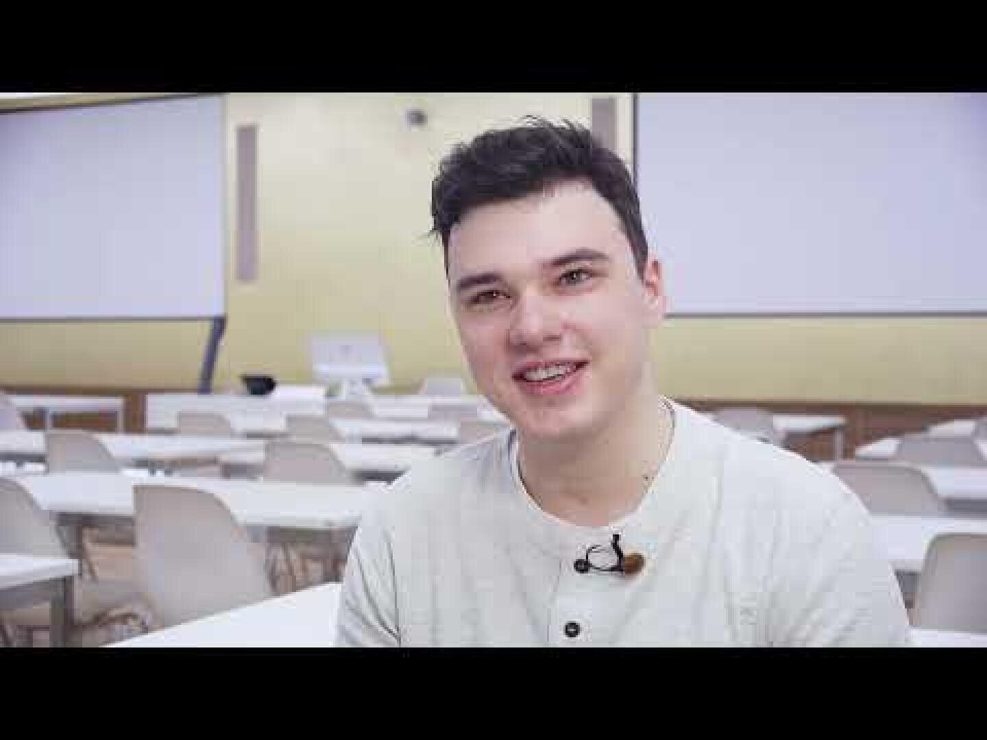 Ein junger Mann mit kurzen dunklen Haaren, der ein helles Hemd und ein kleines Mikrofon trägt, sitzt lächelnd in einem Klassenzimmer mit leeren weißen Tischen und großen Projektionsflächen im Hintergrund.