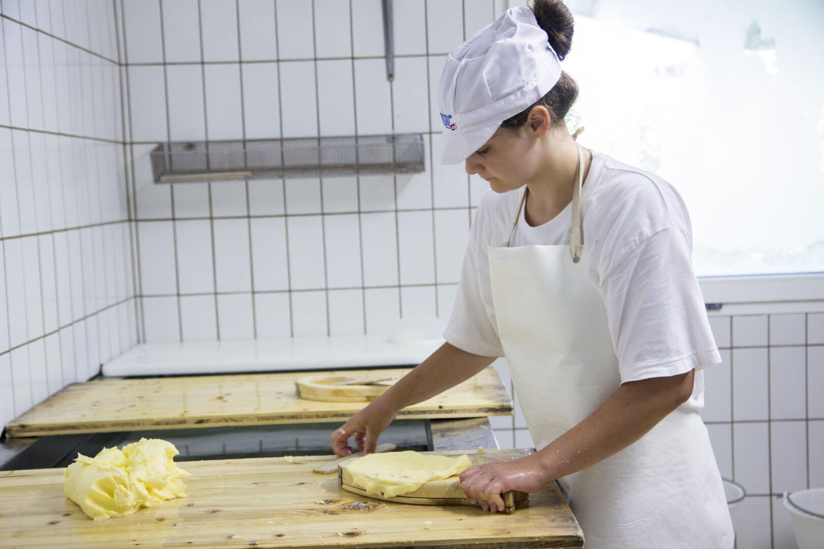 Eine Person mit weißer Schürze und Mütze formt Butterblöcke auf einem Holztisch in einer gekachelten Küche.
