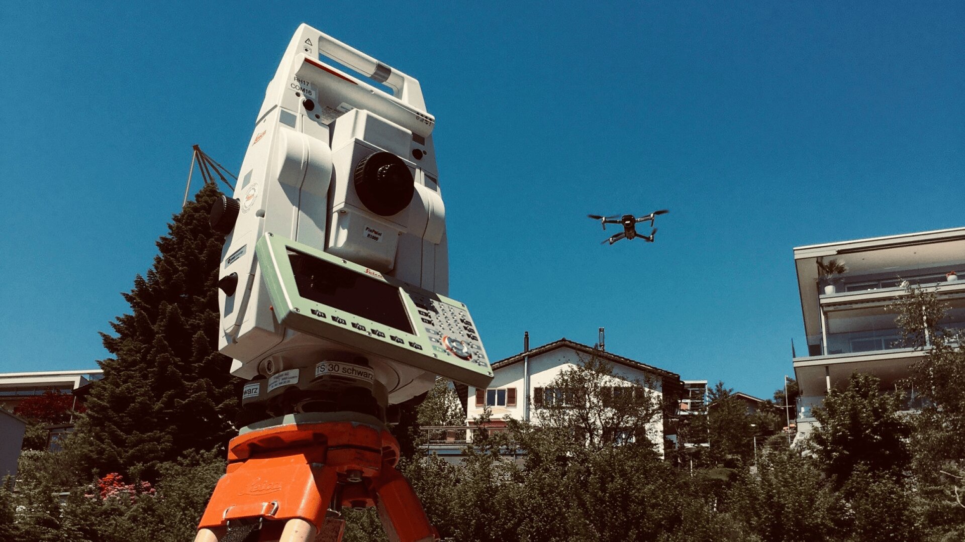 Ein Tachymeter auf einem Stativ steht im Freien, während eine Drohne am blauen Himmel über Häusern und Bäumen im Hintergrund fliegt.