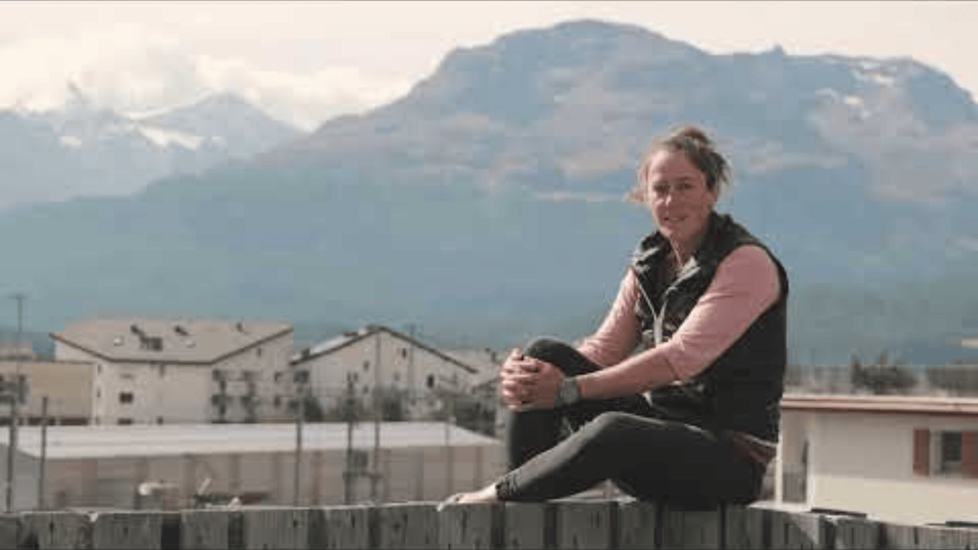 Eine Person sitzt auf einem Holzzaun und lächelt in die Kamera, während im Hintergrund Gebäude und hohe Berge unter einem bewölkten Himmel zu sehen sind.