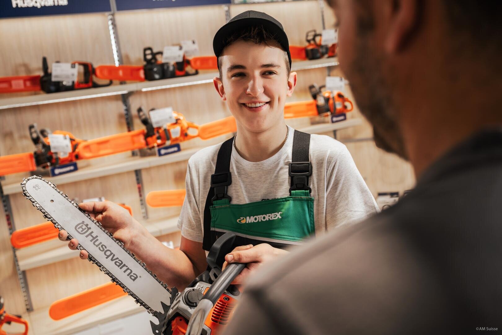 Ein junger Angestellter in grüner Latzhose und Mütze lächelt, während er eine Husqvarna-Kettensäge in der Hand hält und sie einem Kunden in einem Geschäft zeigt, in dem im Hintergrund Kettensägen in Regalen ausgestellt sind.