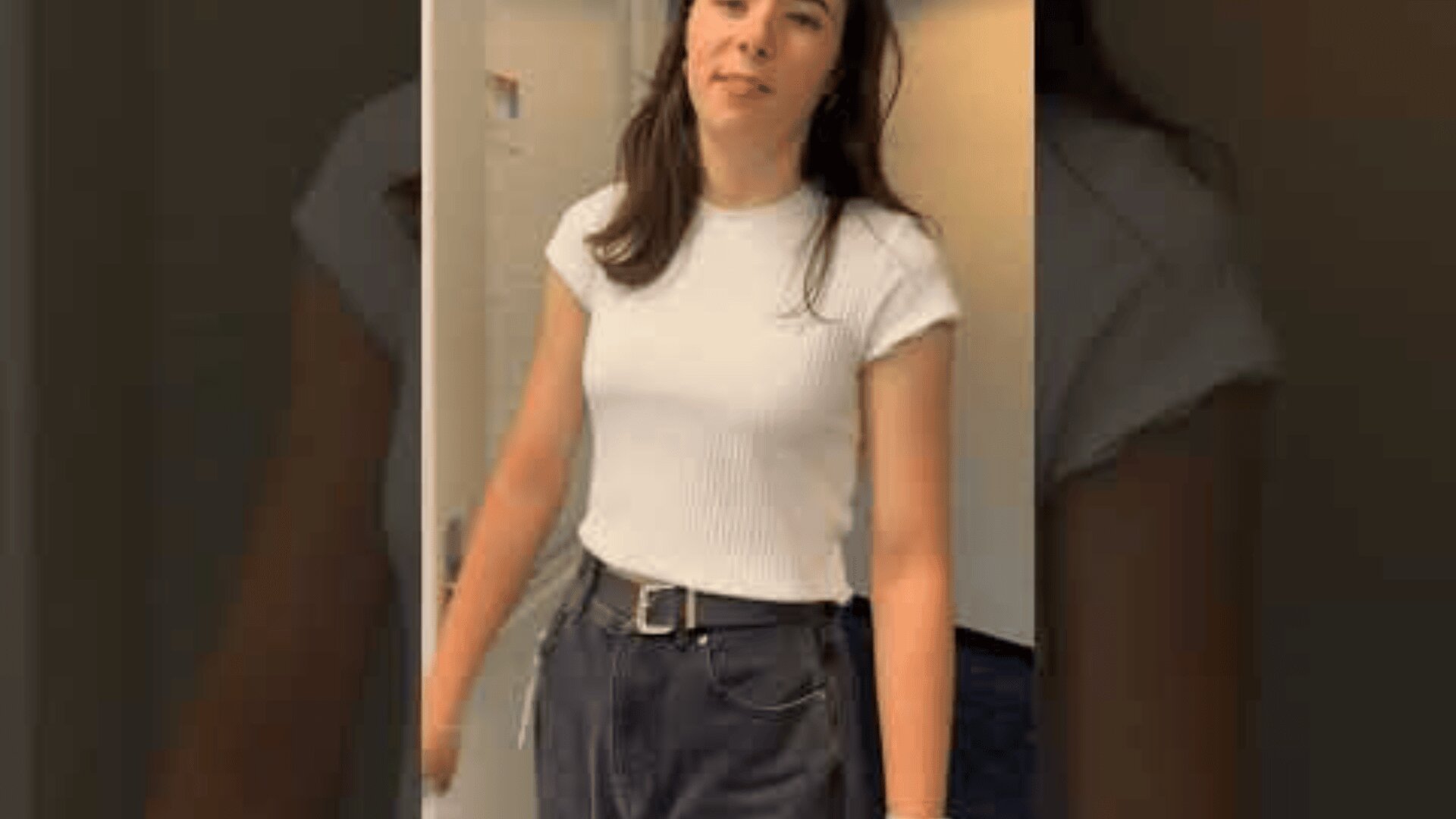Eine junge Frau mit langen braunen Haaren, die ein weißes, kurzärmeliges Oberteil und dunkle Jeans mit hoher Taille trägt, steht in einem Haus und schaut in die Kamera.