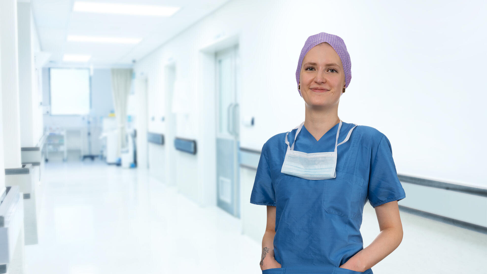 Eine medizinische Fachkraft in blauem Kittel, einer lila OP-Mütze und einer Gesichtsmaske um den Hals steht in einem hellen, modernen Krankenhausflur und lächelt selbstbewusst in die Kamera.