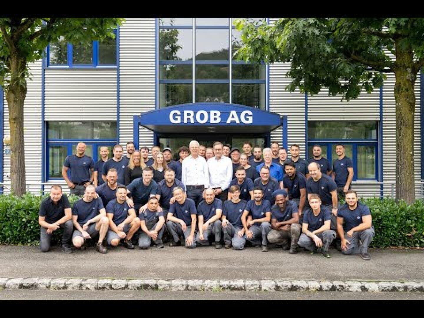 Eine Gruppe von etwa 40 Personen, meist in passenden marineblauen Hemden, steht und kniet vor einem Gebäude mit einem Schild der GROB AG über dem Eingang, umgeben von Bäumen und Hecken.