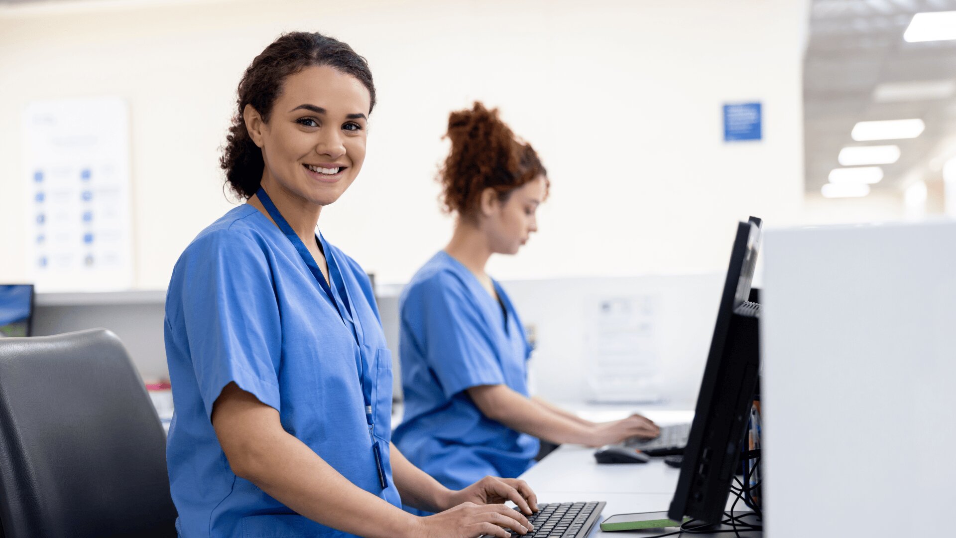 Zwei weibliche medizinische Fachkräfte in blauen Kitteln arbeiten an Computern in einem hellen, modernen medizinischen Büro. Die eine lächelt in die Kamera, während sie tippt, und die andere ist auf ihren Bildschirm im Hintergrund konzentriert.
