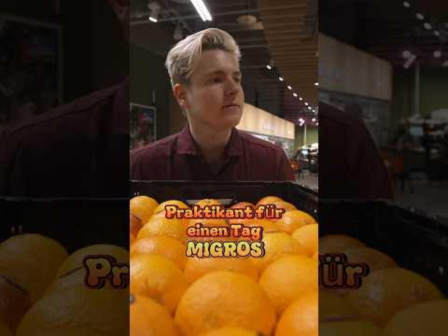 Eine Person mit kurzem blondem Haar steht in einem Supermarkt hinter einer Kiste mit Orangen. Über den Orangen steht in fetter deutscher Schrift: Praktikant für einen Tag MIGROS. Im Hintergrund des Ladens sind Regale und Produkte zu sehen.
