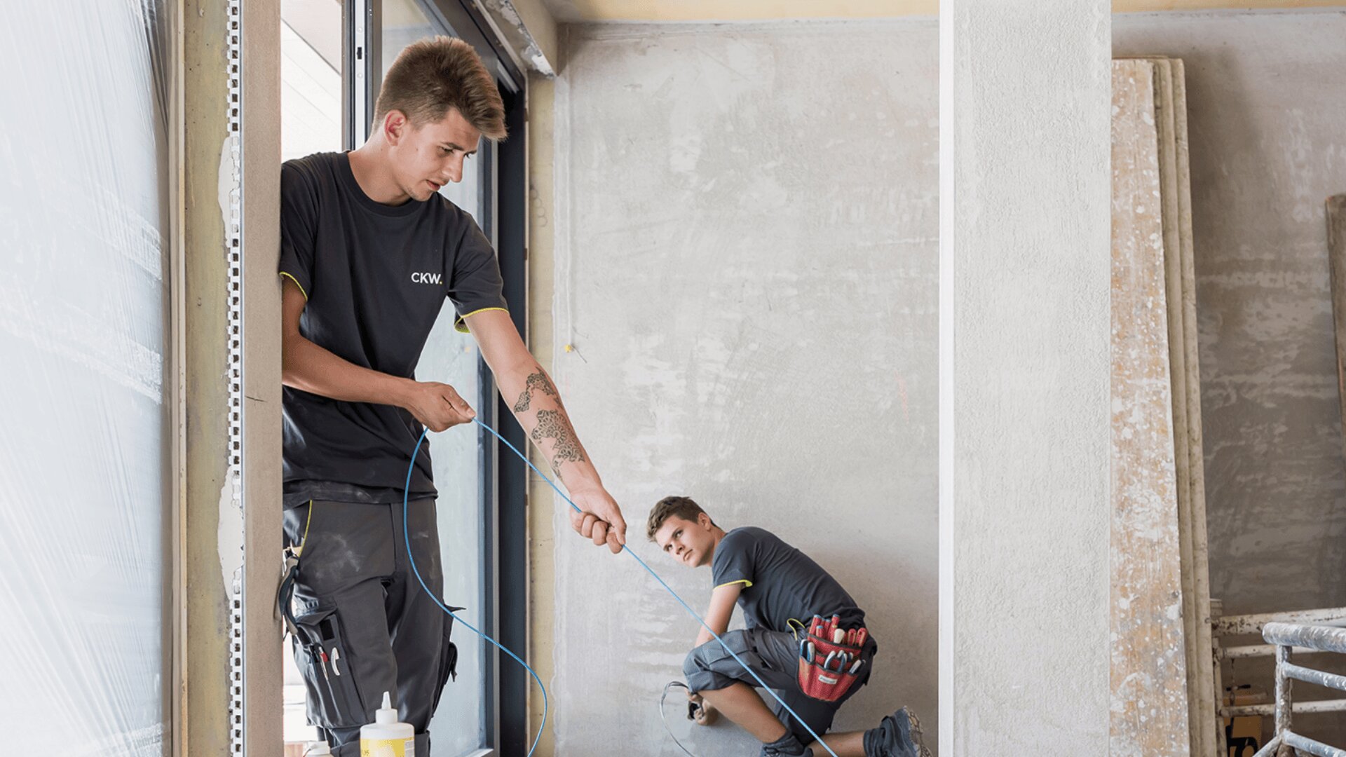 Zwei Bauarbeiter, ein stehender und ein kniender, arbeiten mit blauen Kabeln auf einer Baustelle. Der stehende Arbeiter, der ein graues Hemd mit CKW-Logo trägt, hält das Kabel, während der kniende Arbeiter, der einen Werkzeuggürtel trägt, daran zieht.