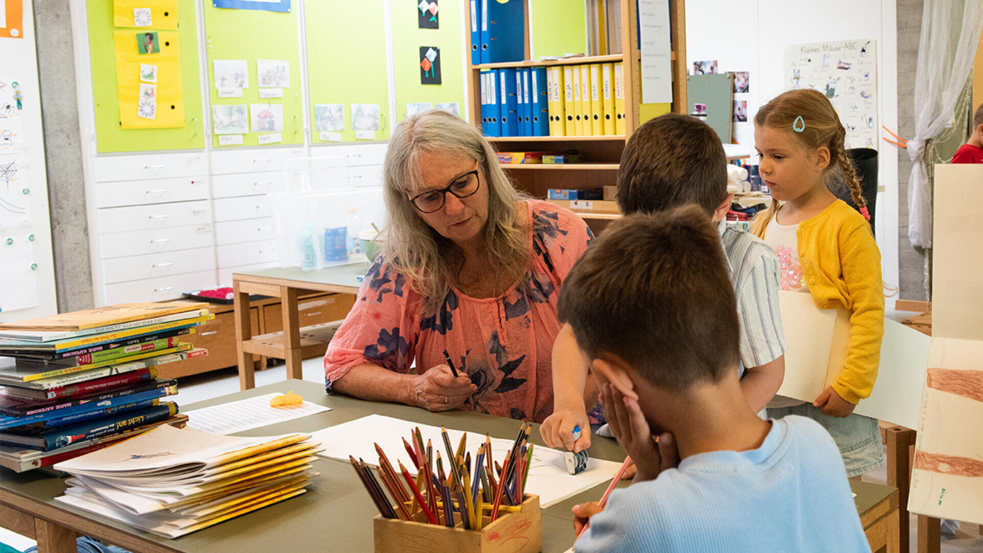 Eine Lehrerin mit grauem Haar sitzt an einem Tisch und hilft jungen Schülern in einem hellen Klassenzimmer, das mit bunten Mappen, Zeichnungen und Kunstzubehör gefüllt ist. Die Kinder sind um sie herum versammelt, einige halten Papiere in der Hand.