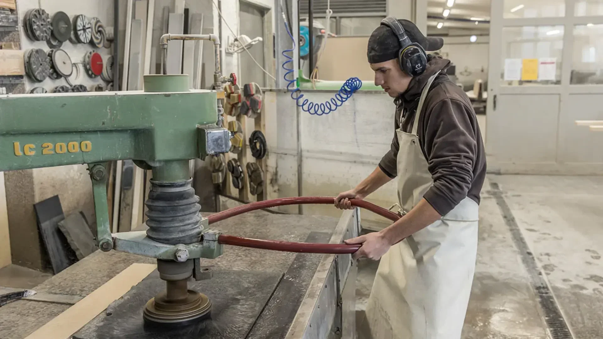 Ein Arbeiter mit Schürze und Gehörschutz bedient eine große industrielle Poliermaschine in einer Werkstatt und führt einen roten Schlauch, während er die Oberfläche einer Steinarbeitsplatte glättet.