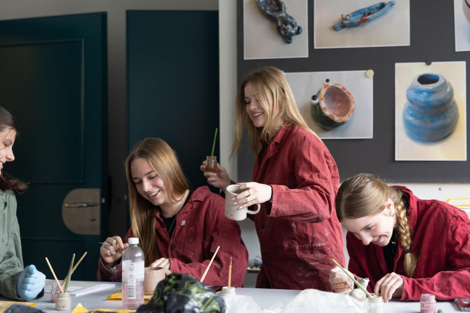 Drei junge Frauen in roten Kitteln lächeln und lachen, während sie an einem Tisch Töpferwaren bemalen. Vor ihnen liegen Pinsel und Zubehör, hinter ihnen hängen Keramikkunstwerke an der Wand.