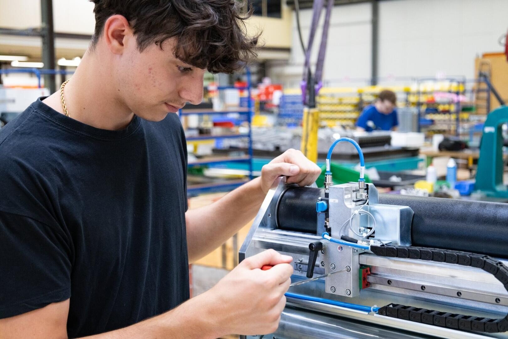 Ein junger Mann in einem schwarzen Hemd bedient Maschinen in einer hellen Industriewerkstatt, stellt Bedienelemente ein und konzentriert sich auf seine Aufgabe. Im Hintergrund sind Regale, Werkzeuge und ein weiterer Arbeiter zu sehen.