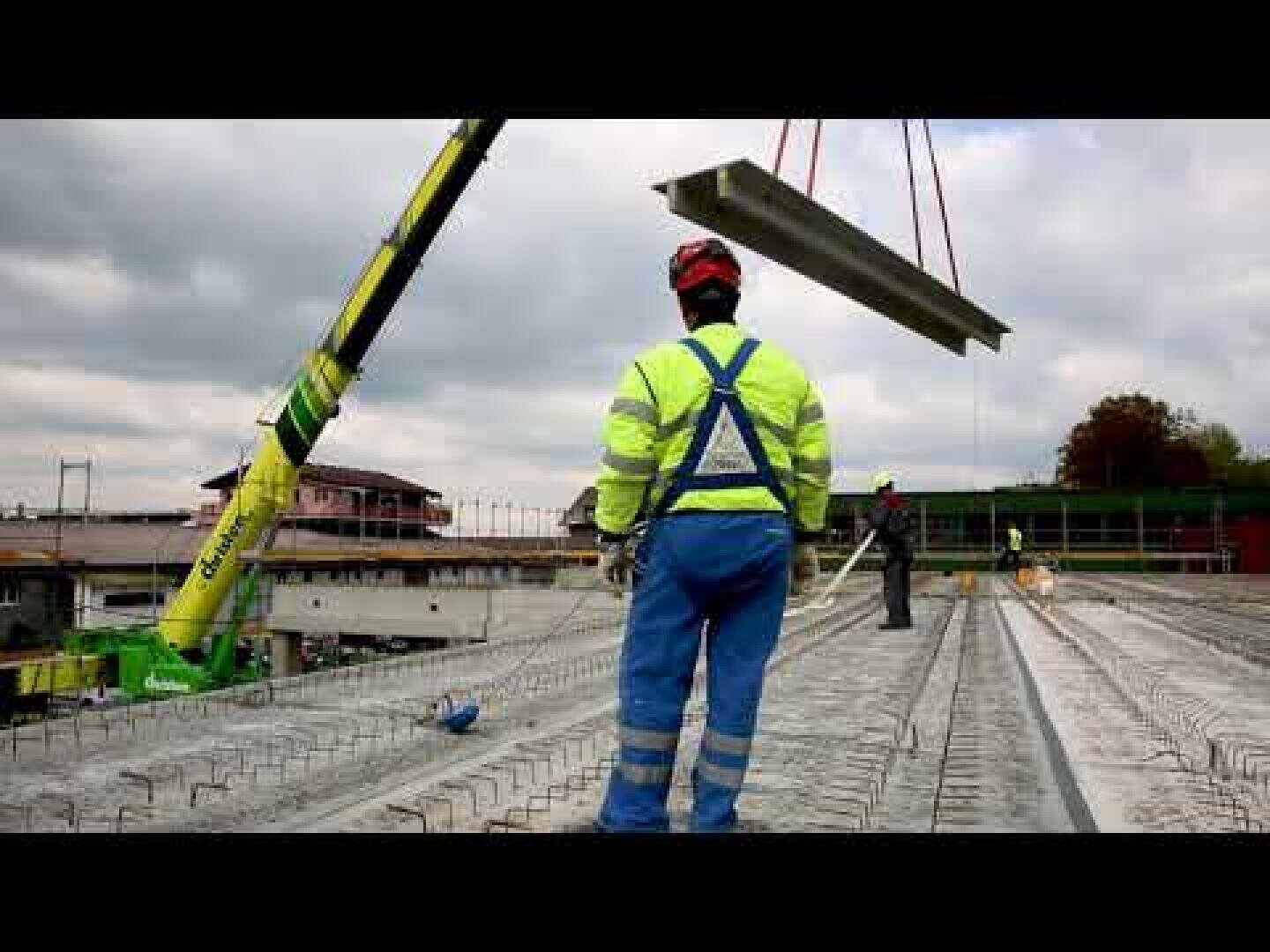 Ein Bauarbeiter in Warnweste und Helm steht auf einer Baustelle und beobachtet, wie ein Kran bei bewölktem Himmel eine große Betonplatte anhebt. Im Hintergrund sind andere Arbeiter und Baumaterialien zu sehen.