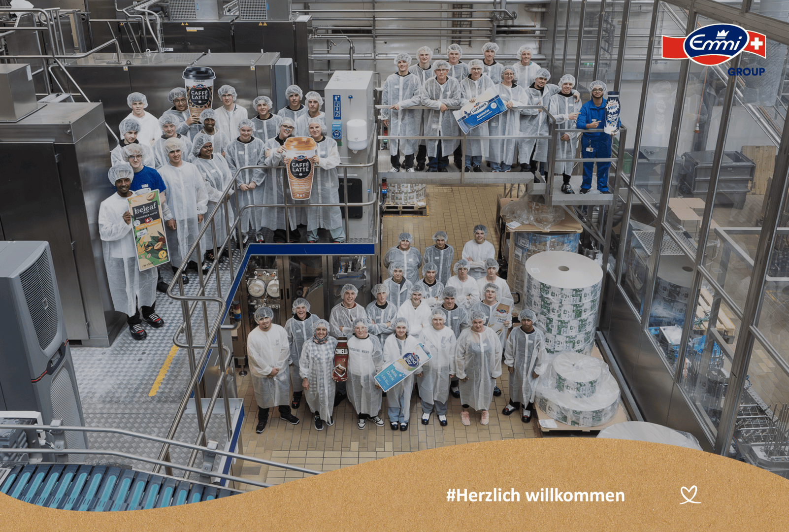 Eine große Gruppe von Menschen in weißen Laborkitteln und Haarnetzen steht in einem Lebensmittelproduktionsbetrieb zusammen und lächelt in die Kamera. Auf dem Bild ist das Logo der Emmi Gruppe und Herzlich willkommen zu sehen.