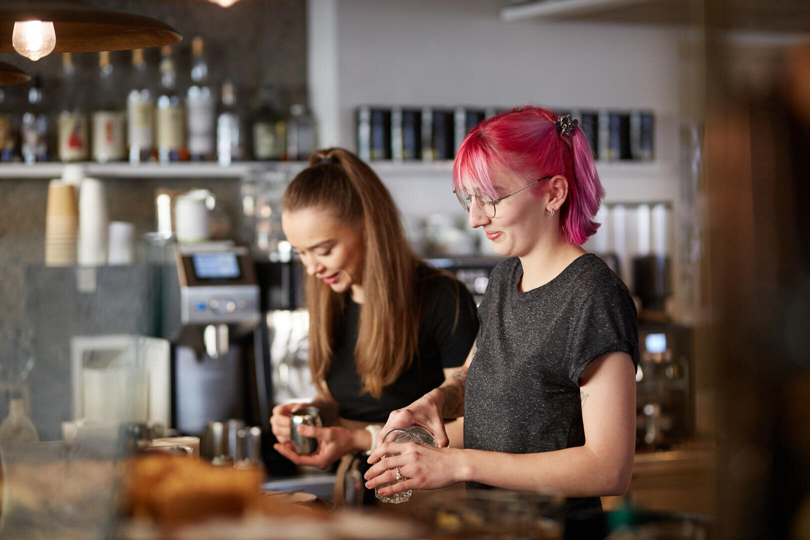 Zwei Baristas arbeiten hinter der Theke eines Cafés, die eine mit langen braunen Haaren, die andere mit leuchtend pinkfarbenen Haaren; beide bereiten mit konzentriertem Gesichtsausdruck Getränke zu. Im Hintergrund sind Regale mit Flaschen und Tassen zu sehen.