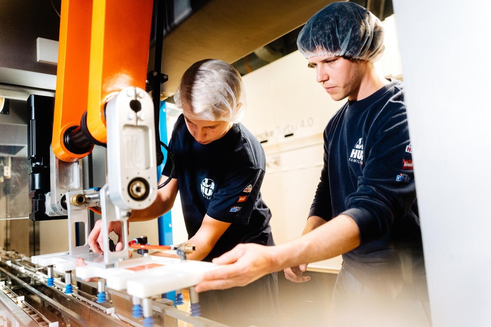 Zwei Arbeiter, die ein Haarnetz und ein dunkles Hemd tragen, bedienen oder überprüfen eine Maschine mit orangefarbenen und weißen Bauteilen in einer Fabrik oder in der Industrie und konzentrieren sich dabei ganz auf ihre Aufgabe.
