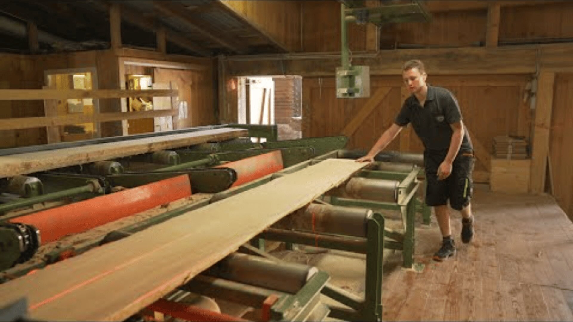 Eine Person bedient eine große grüne und rote Holzbearbeitungsmaschine, die ein langes Holzbrett durch Rollen in einer Werkstatt mit Holzwänden und -böden führt.