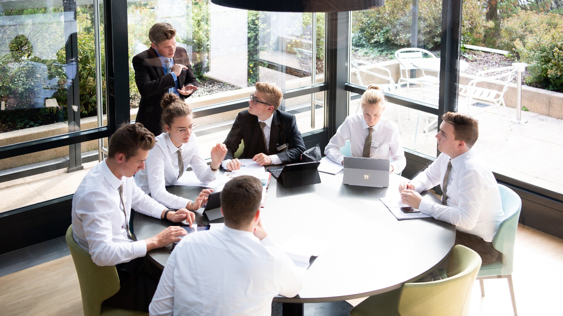 Sechs Personen in Geschäftskleidung sitzen um einen runden Tisch in einem modernen, sonnendurchfluteten Konferenzraum, arbeiten mit Laptops und machen sich Notizen, während eine Person aufsteht und spricht, so als würde sie die Sitzung leiten.