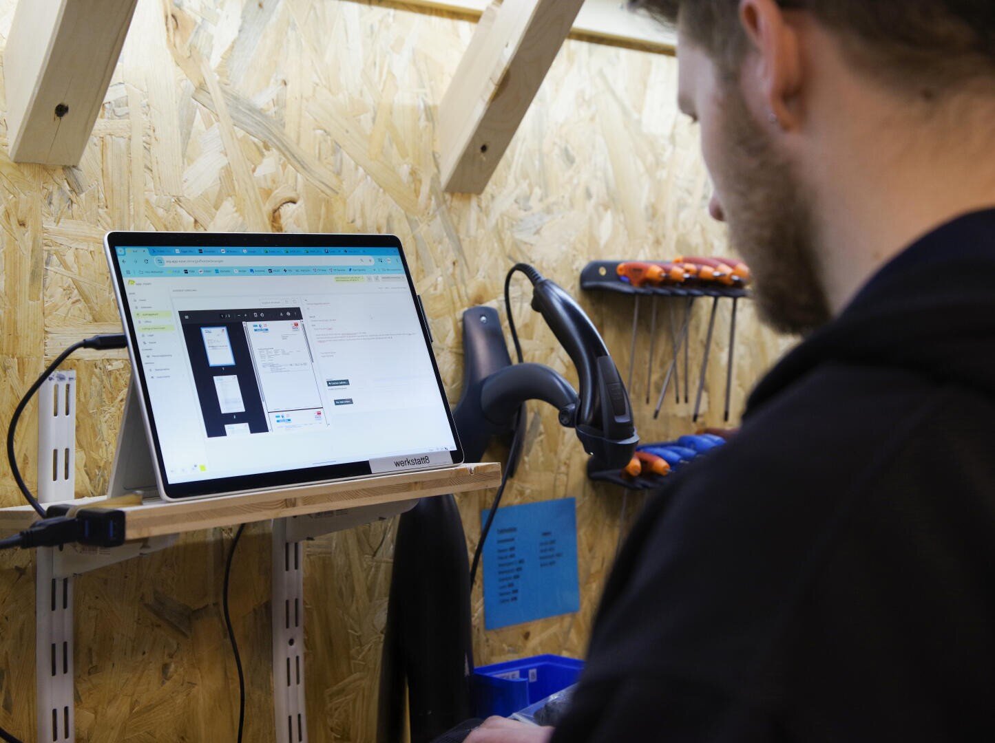 Eine Person verwendet einen Computer an einer Werkbank mit Werkzeugen, einem Barcode-Scanner und einem an der Wand montierten Monitor, auf dem eine Softwareoberfläche in einer Werkstattumgebung angezeigt wird.