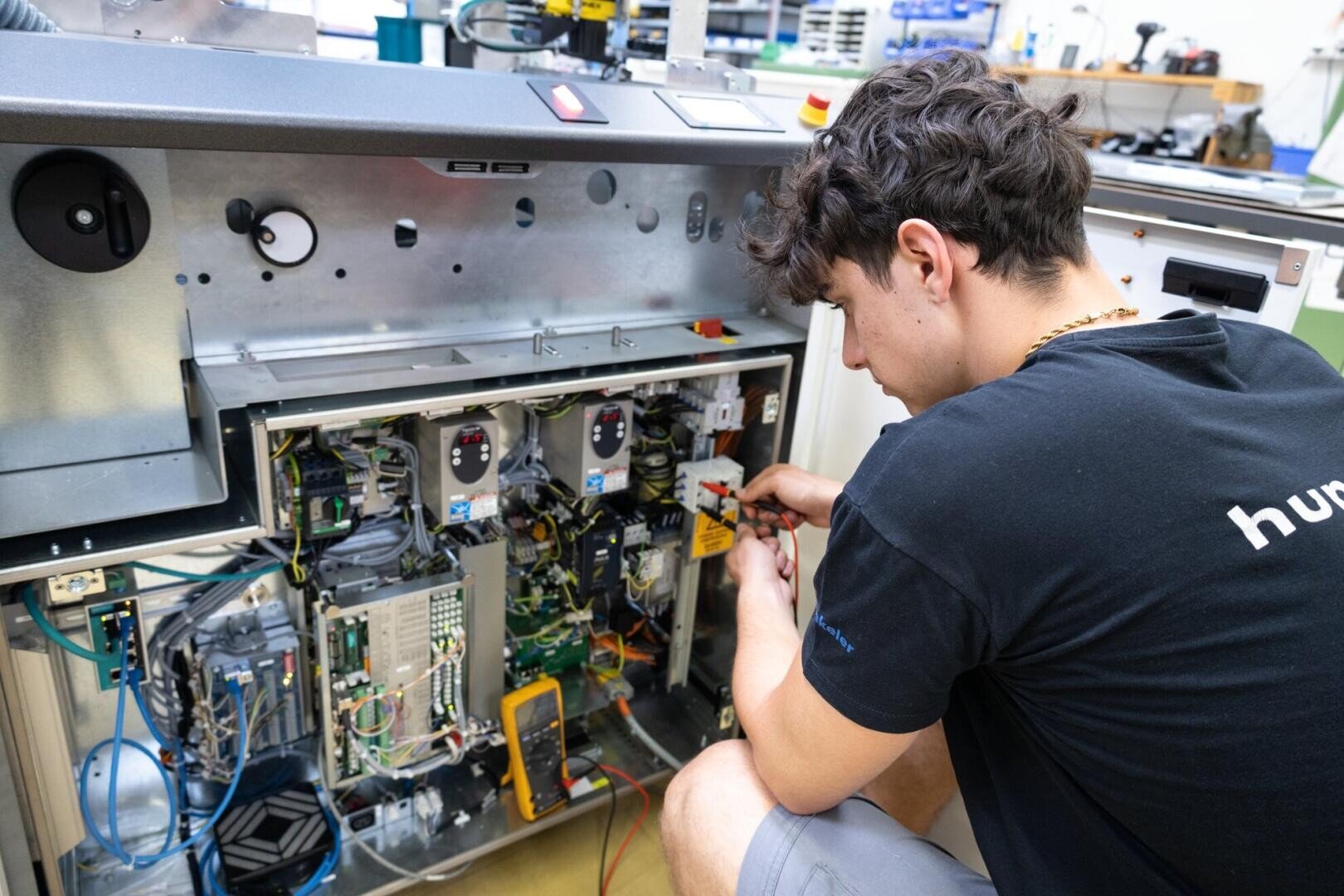 Eine Person in einem schwarzen Hemd arbeitet mit Werkzeugen an der Verkabelung einer offenen elektronischen Industriemaschine in einer Werkstatt.