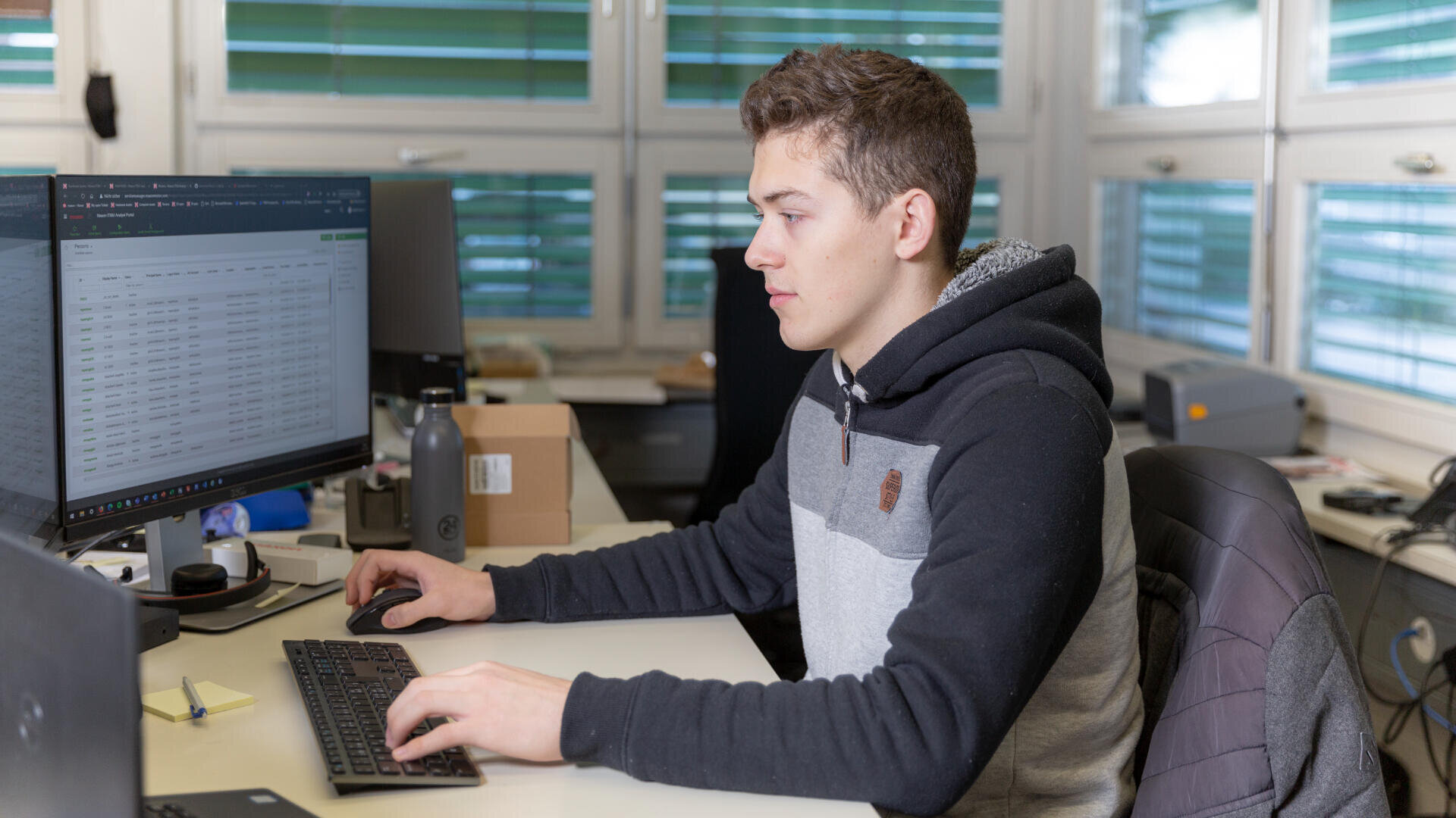 Ein junger Mensch mit Kapuzenpulli arbeitet an einem Schreibtisch mit zwei Computermonitoren und tippt auf einer Tastatur in einem modernen Büro mit großen Fenstern im Hintergrund.