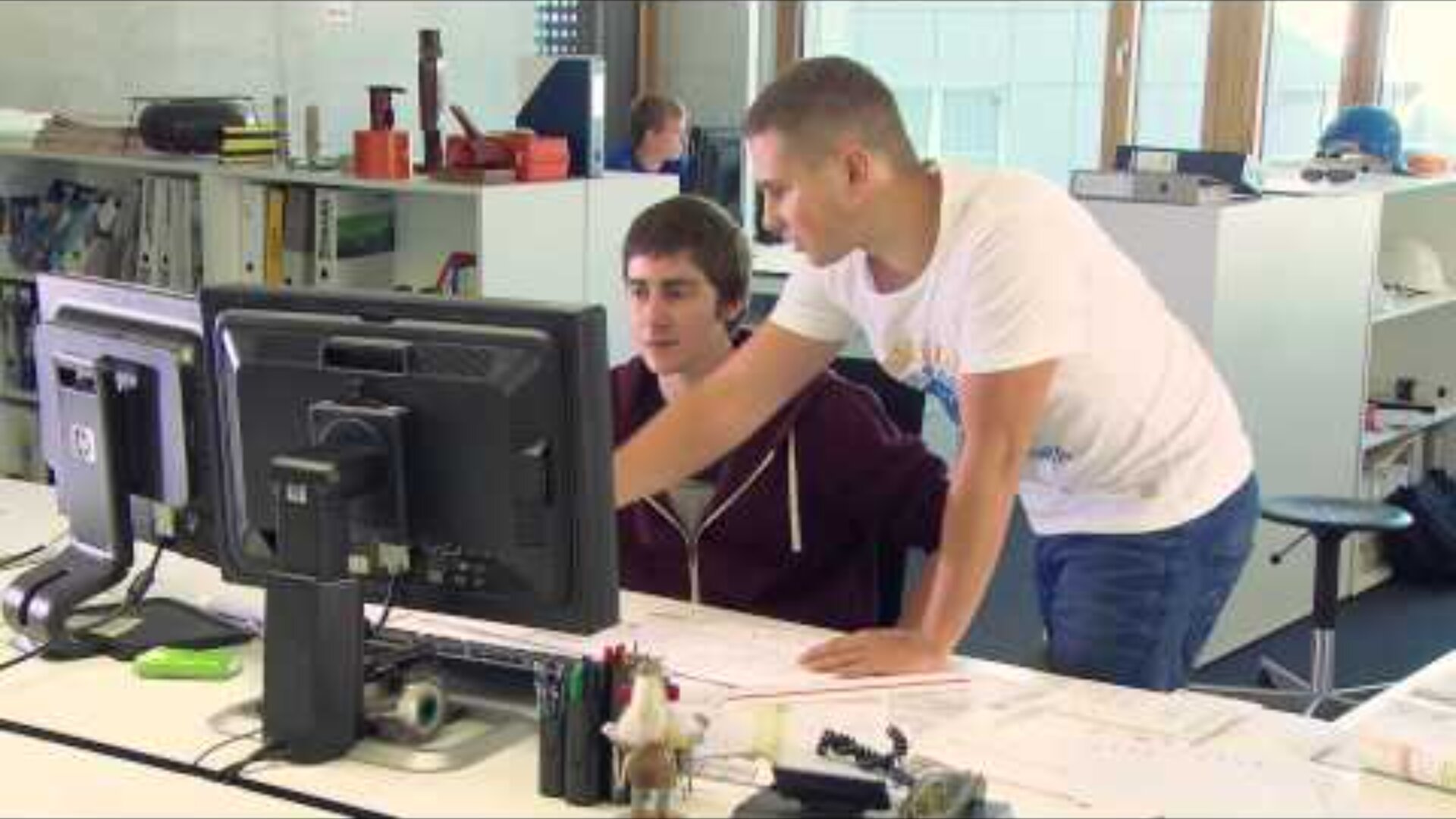 Zwei Personen arbeiten in einem modernen Büro an einem Schreibtisch zusammen und schauen auf Computerbildschirme. Eine Person sitzt, die andere steht und zeigt auf den Bildschirm, beide sind auf ihre Aufgabe konzentriert. Auf dem Schreibtisch sind Büromaterialien und Papierkram zu sehen.