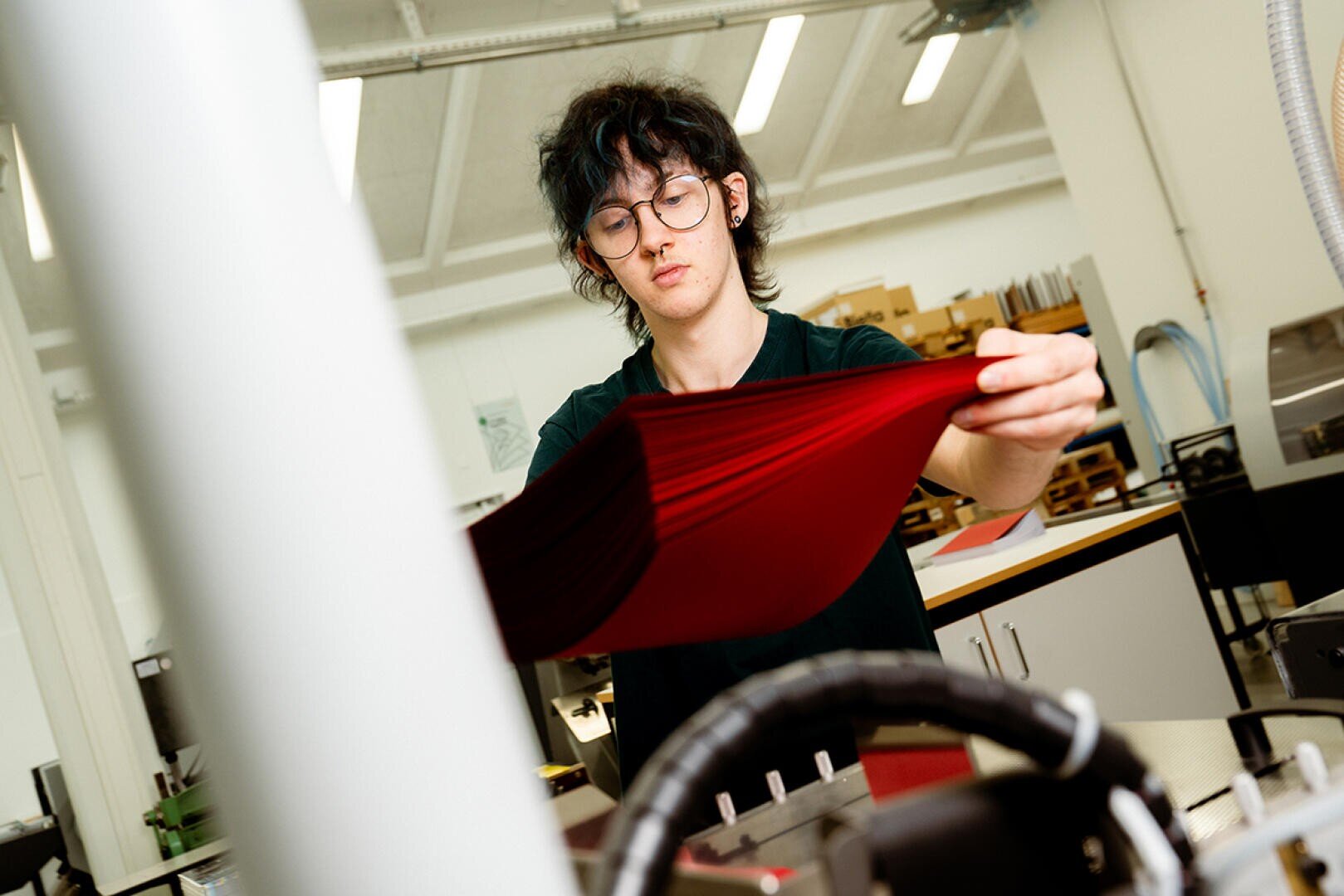 Eine Person mit Brille und dunklem Haar prüft einen großen Stapel roter Papierbögen in einem hell erleuchteten, modernen Arbeitsraum mit Regalen und Geräten im Hintergrund.