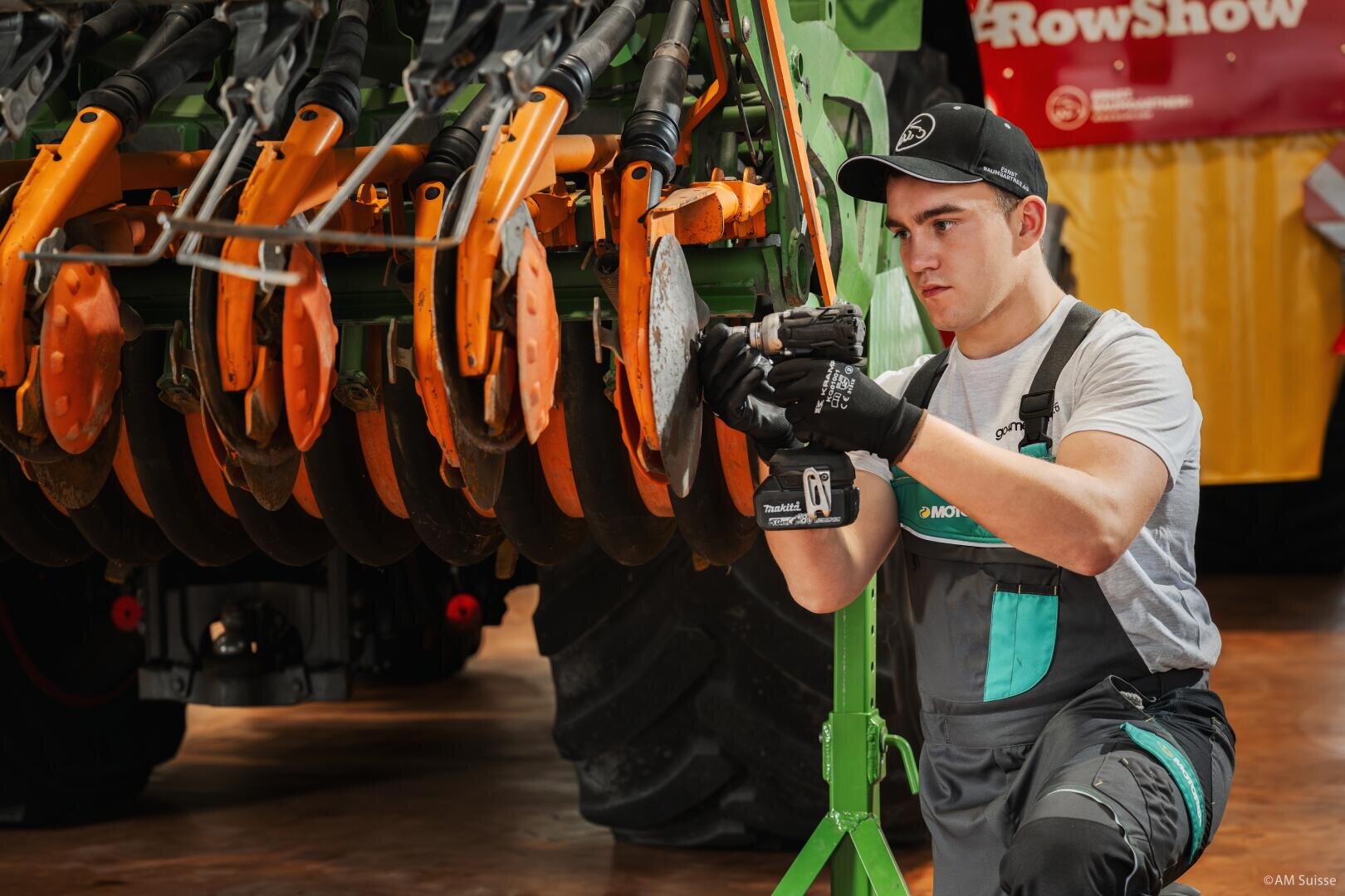 Ein junger Mann in Arbeitsoverall und Handschuhen kniet neben einer großen Landmaschine und benutzt ein Elektrowerkzeug, um orangefarbene Metallscheiben einzustellen oder zu reparieren. Die Innenaufnahme lässt auf eine Werkstatt oder eine Gerätevorführung schließen.
