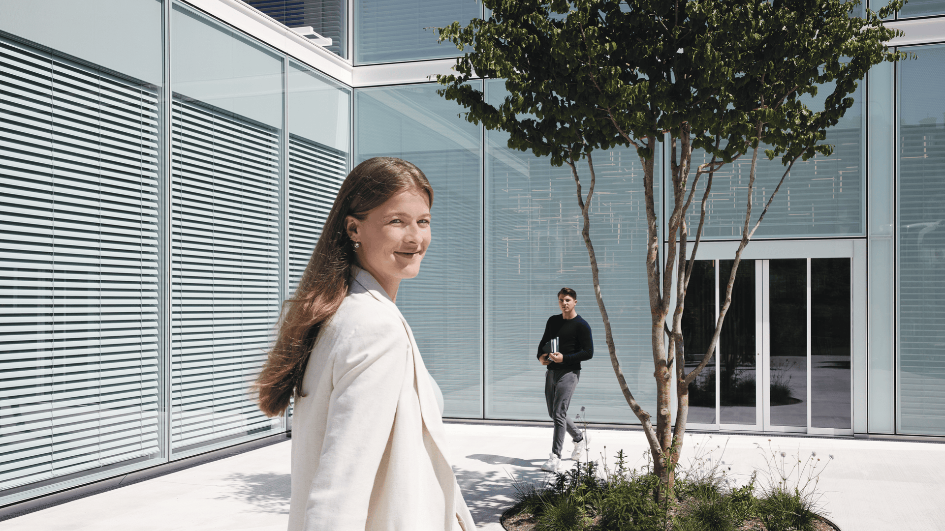 Eine Frau in einem weißen Blazer lächelt, während sie vor einem modernen Gebäude mit großen Glasfenstern spazieren geht. Ein Mann in dunkler Kleidung geht im Hintergrund in der Nähe eines kleinen Baumes und von Glastüren. Die Szene ist hell und sonnig.