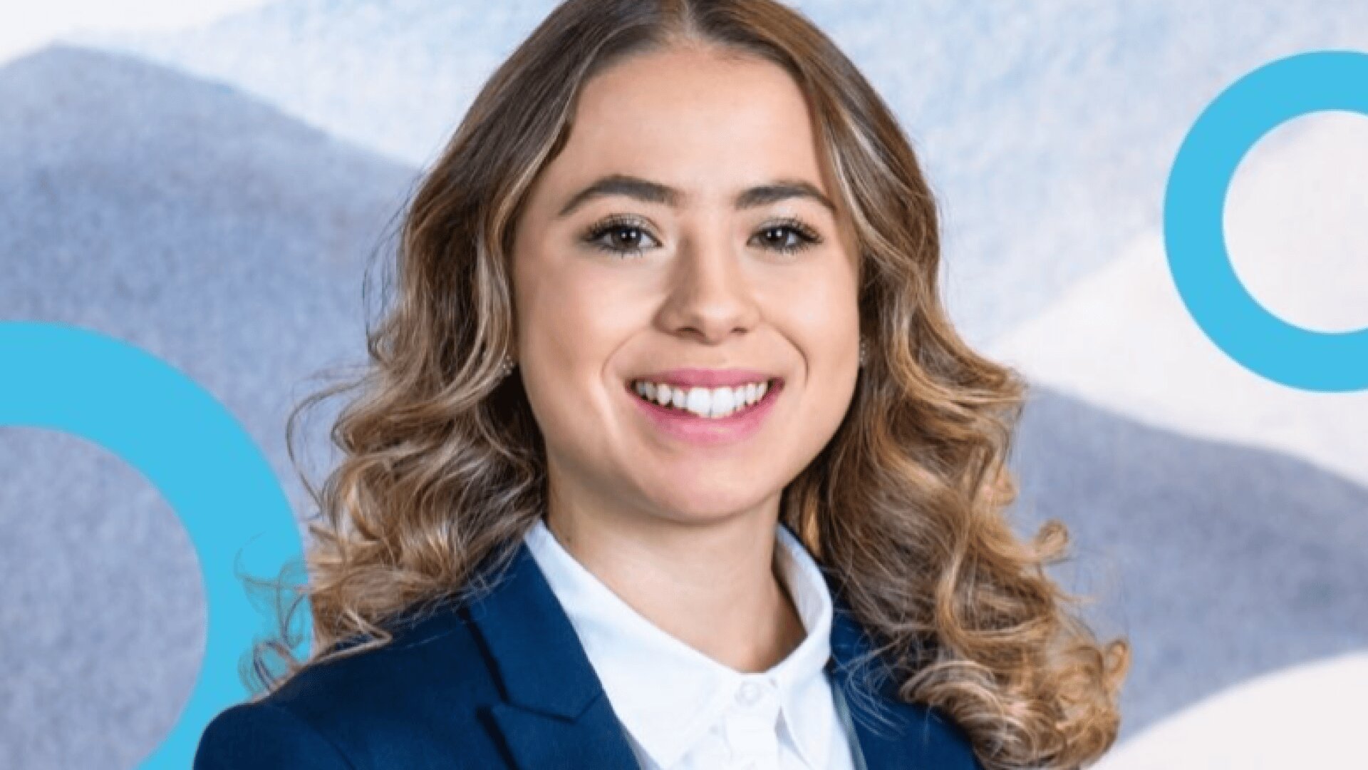 Eine Frau mit langen, lockigen hellbraunen Haaren lächelt. Sie trägt einen dunkelblauen Blazer und ein weißes Hemd mit Kragen. Im Hintergrund sind abstrakte blaue und weiße Formen und Kreise zu sehen.
