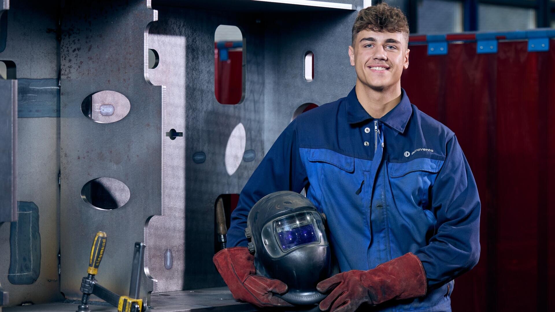 Ein lächelnder Schweißer in blauer Arbeitskleidung, der einen Schweißhelm in der Hand hält und rote Schutzhandschuhe trägt, in einer Werkstatt.
