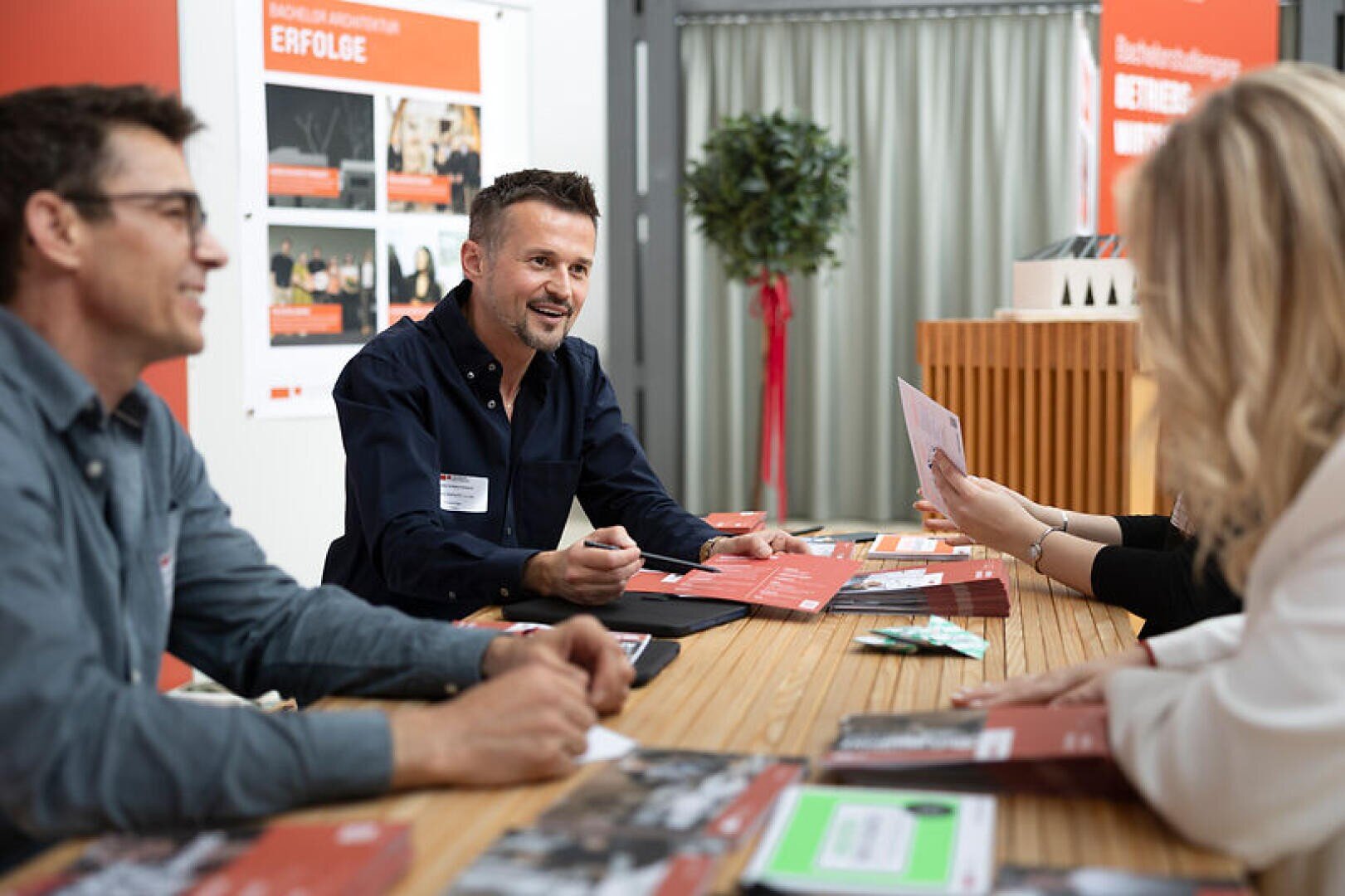 Drei Personen sitzen an einem Tisch mit Broschüren und unterhalten sich mit zwei weiteren Personen, die ihnen gegenüber sitzen. Ein Mann lächelt und hält dabei einen Stift in der Hand. Im Hintergrund sind Plakate und eine Pflanze zu sehen. Die Szene scheint bei einer Veranstaltung in einem Innenraum aufgenommen worden zu sein.