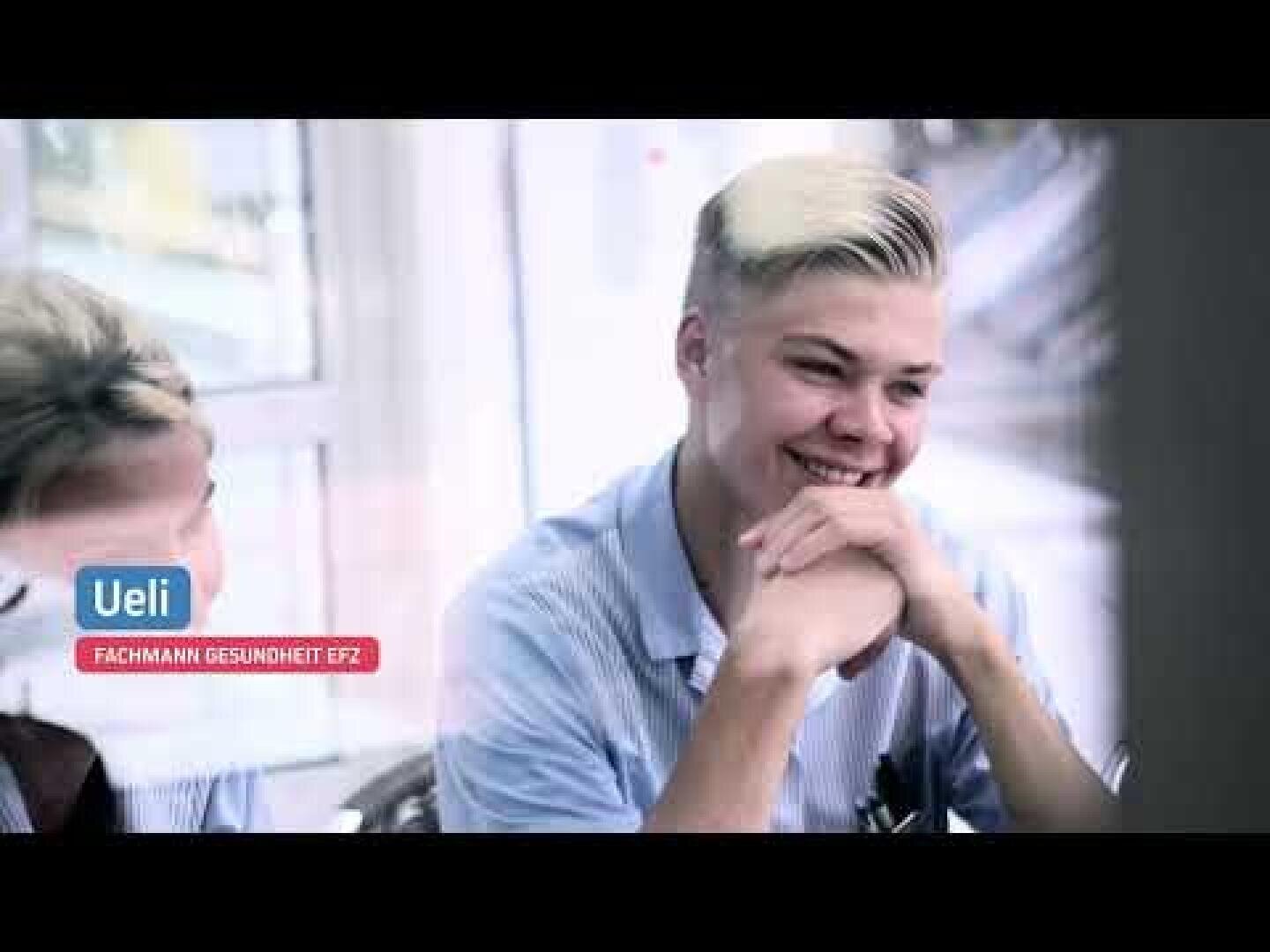 Eine junge Person mit kurzen blonden Haaren und einem gestreiften Hemd sitzt an einem Tisch, lächelt und hält die Hände zusammen. Auf einem Schild steht Ueli, Fachmann Gesundheit EFZ. Neben ihnen ist eine weitere Person teilweise sichtbar.