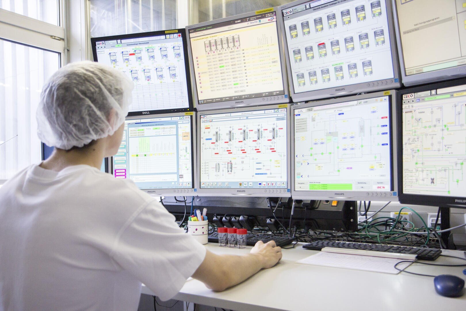 Eine Person, die ein Haarnetz und ein weißes Hemd trägt, überwacht mehrere Bildschirme, auf denen technische Daten und Diagramme in einem Kontrollraum oder Labor angezeigt werden.